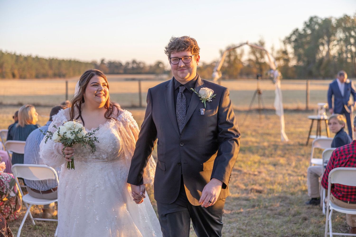A couple walks hand in hand at their outdoor wedding ceremony during golden hour in a rustic field setting.