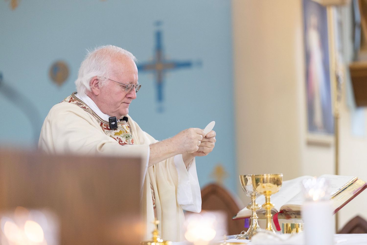 A person in white religious vestments raises a golden chalice during a Catholic mass ceremony.