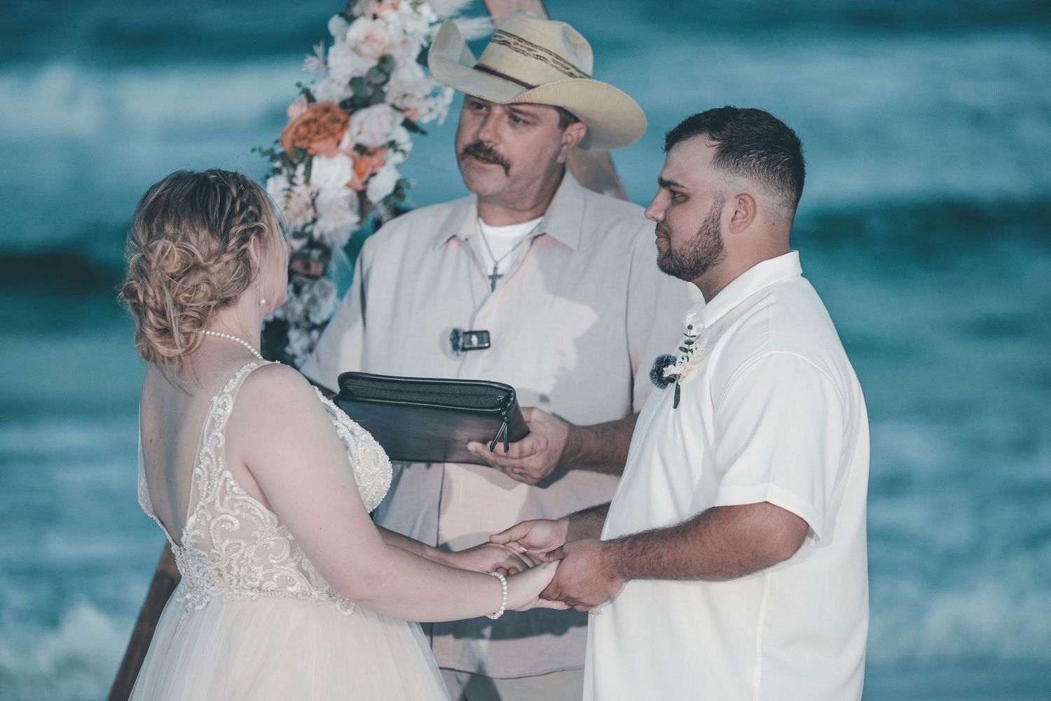 A beachside wedding ceremony takes place with waves crashing in the background on a bright sunny day.