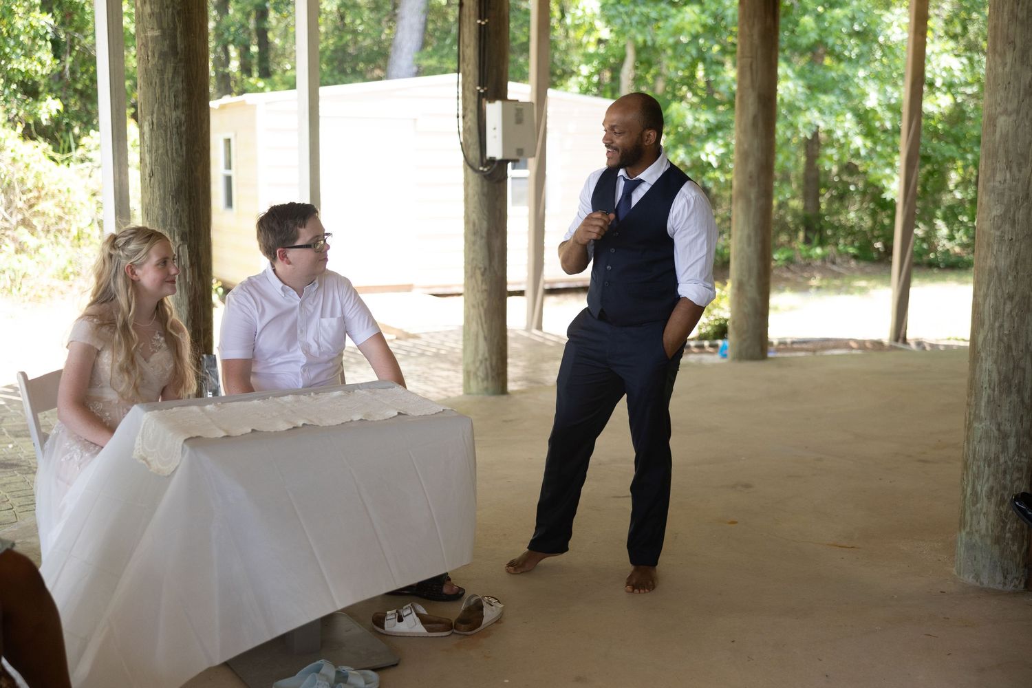 A wedding reception scene under a covered outdoor pavilion with a couple seated at a table while someone speaks.