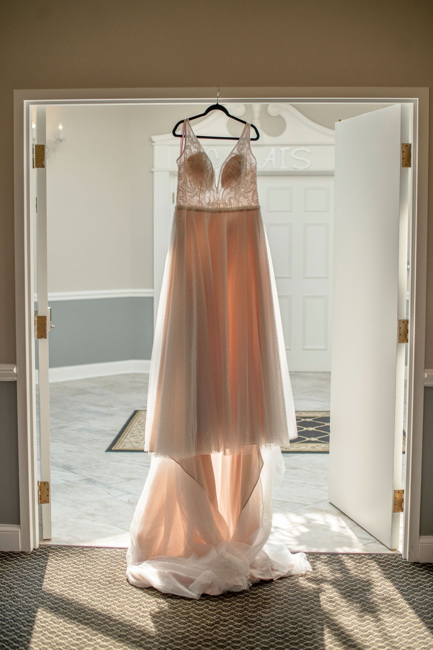 A blush pink wedding dress hangs in front of sunlit doorways showing its delicate silhouette and flowing design.