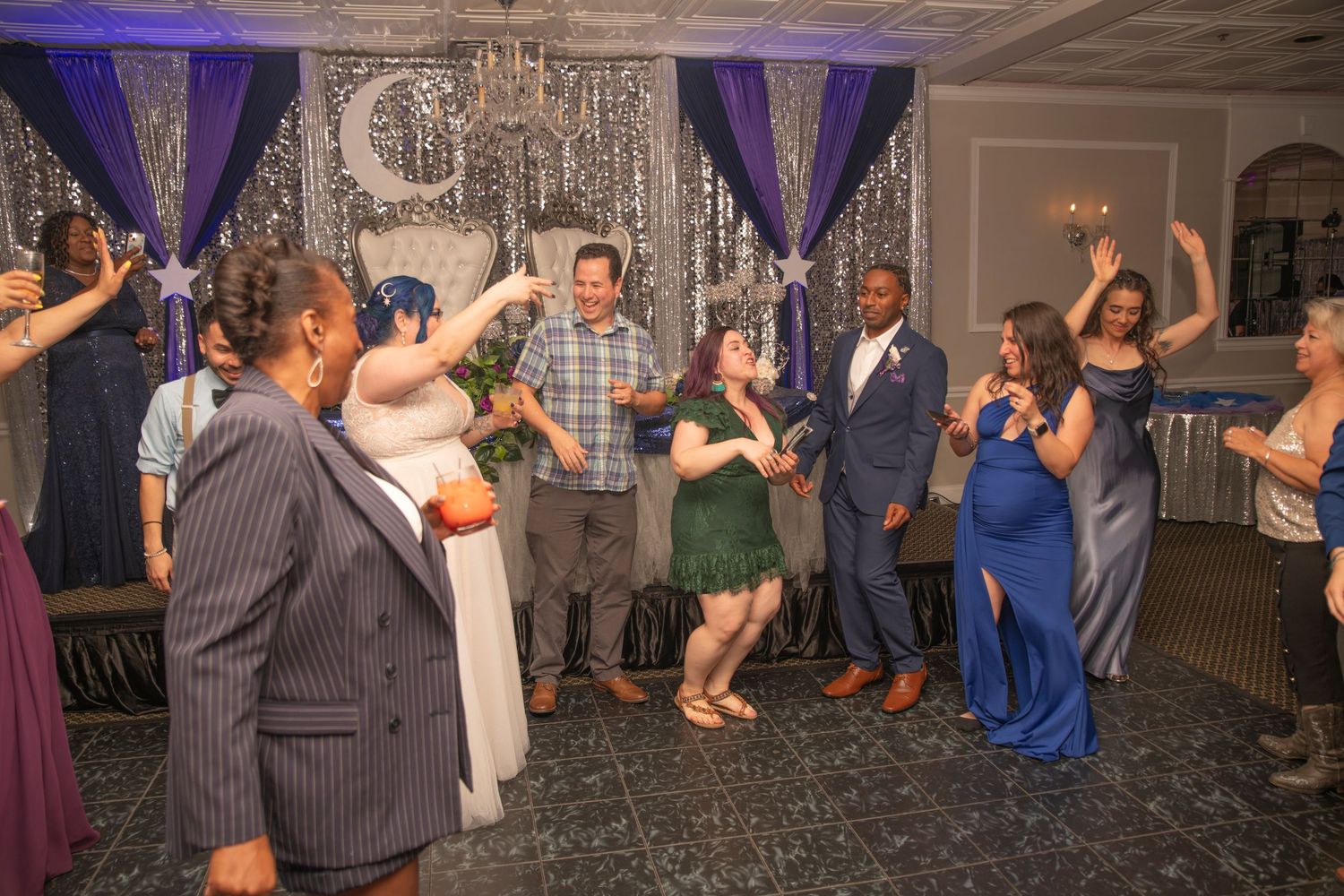 Wedding guests dancing and celebrating on the dance floor under purple decor and silver tinsel at an elegant reception.