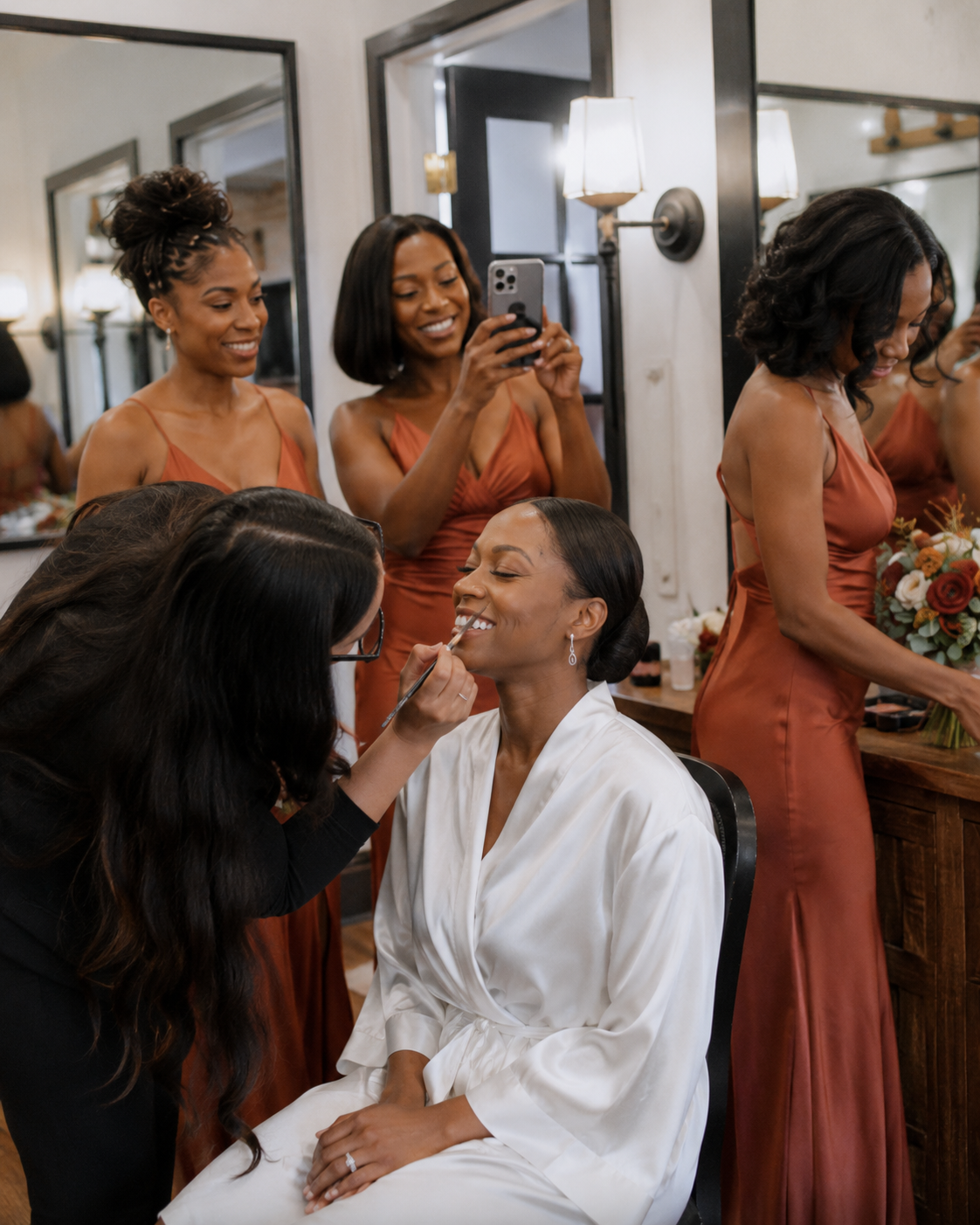 Bride gets makeup done in white robe while smiling bridesmaids in rust-colored dresses assist during wedding preparations.