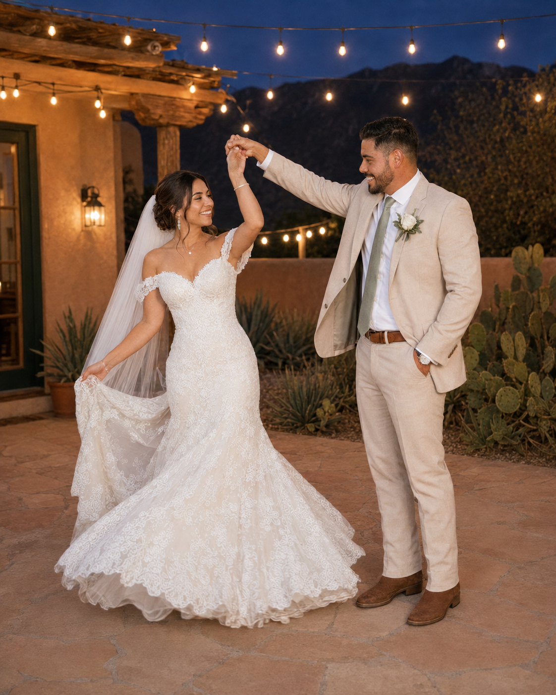 Groom twirls bride in lace mermaid gown under string lights at romantic desert wedding venue at night.