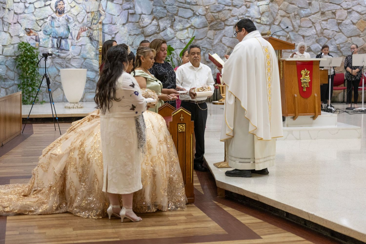 A quinceañera ceremony in a church with a young woman in a gold gown receiving blessings from a priest.