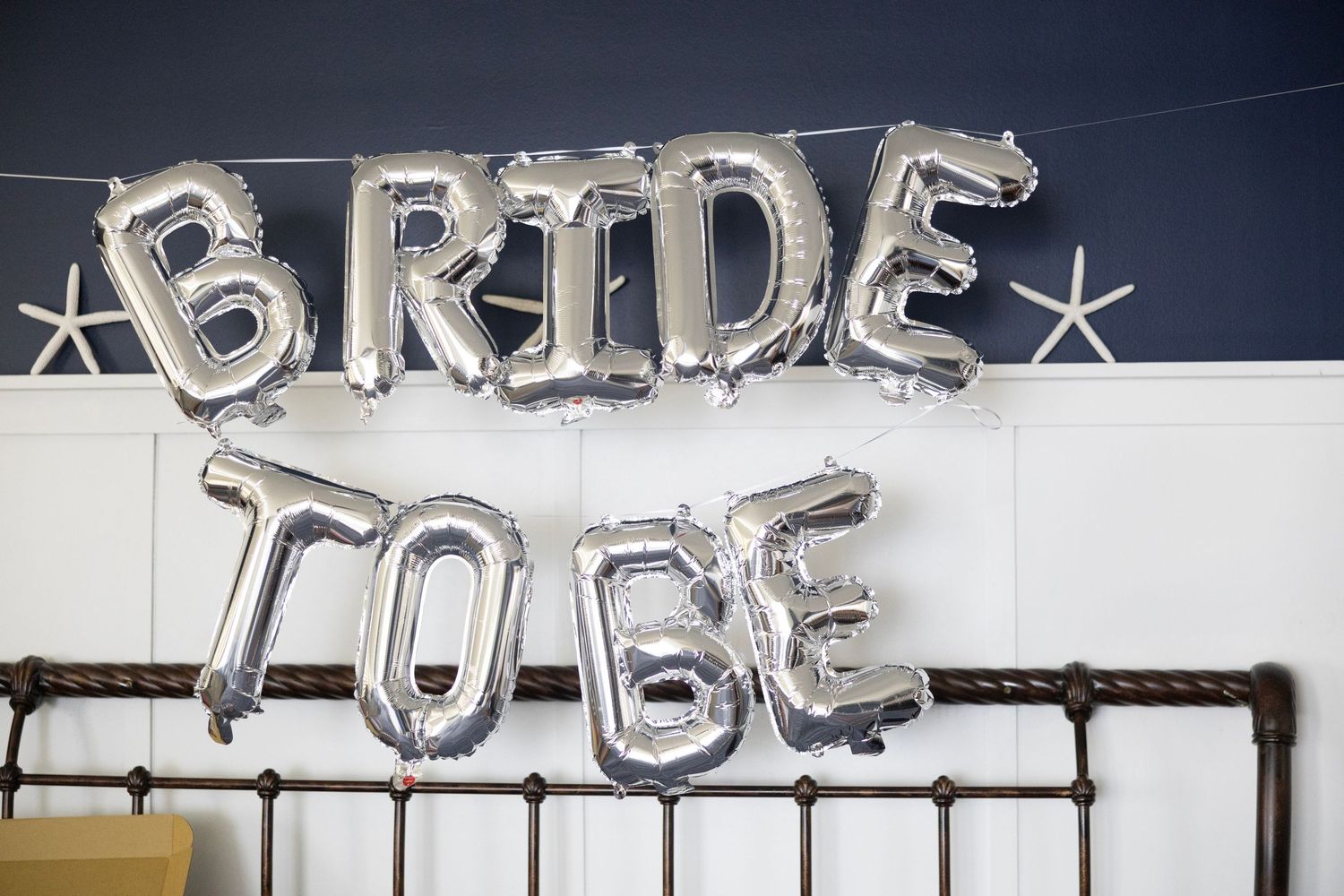 Silver foil balloon letters spelling BRIDE TO BE hung on a dark wall for a bridal shower decoration.