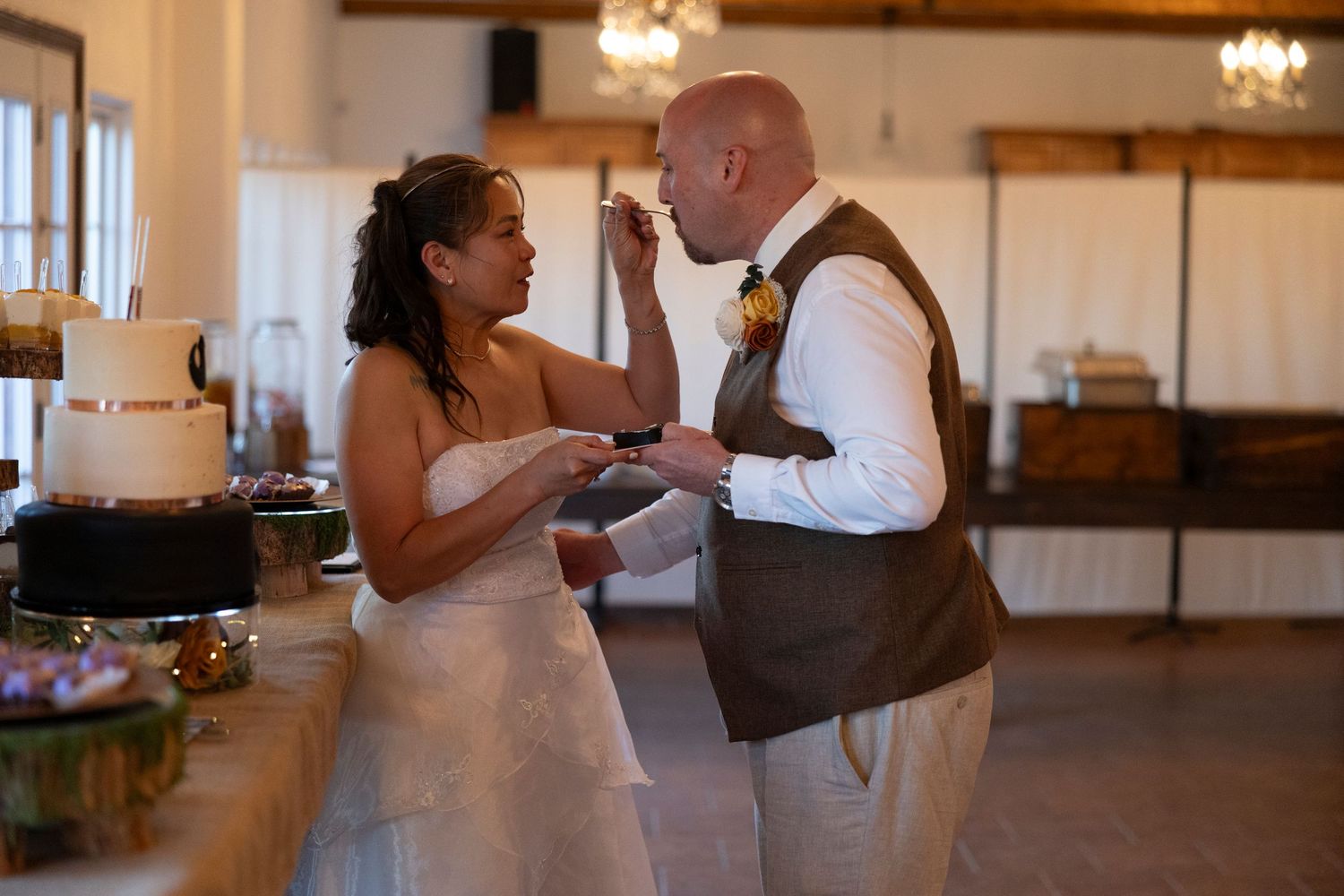 Bride and groom sharing wedding cake at reception, smiling joyfully in elegant venue with tiered cake visible in background.