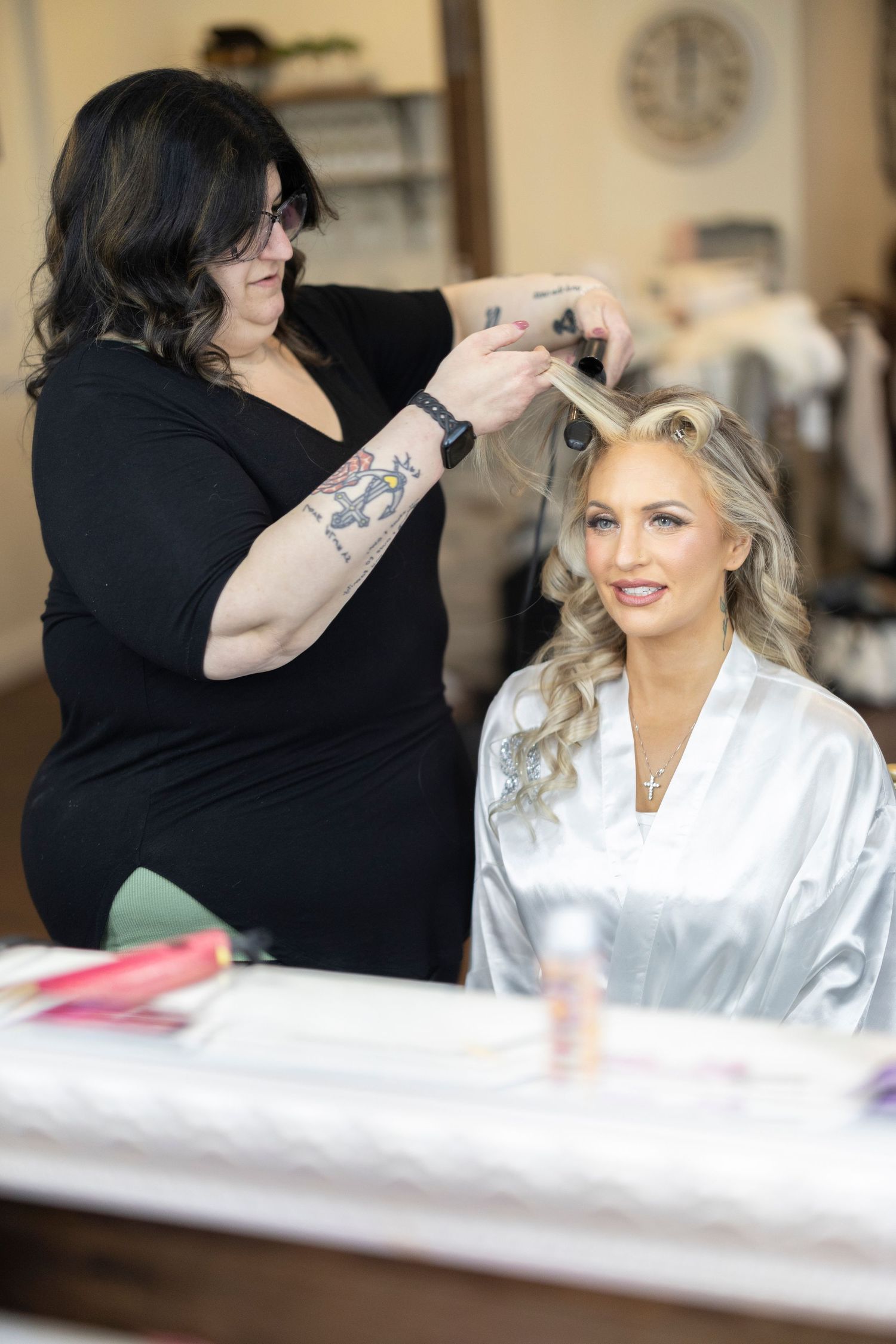 A hairstylist in black attire styles a client's hair in a salon setting while the client wears a white satin robe.