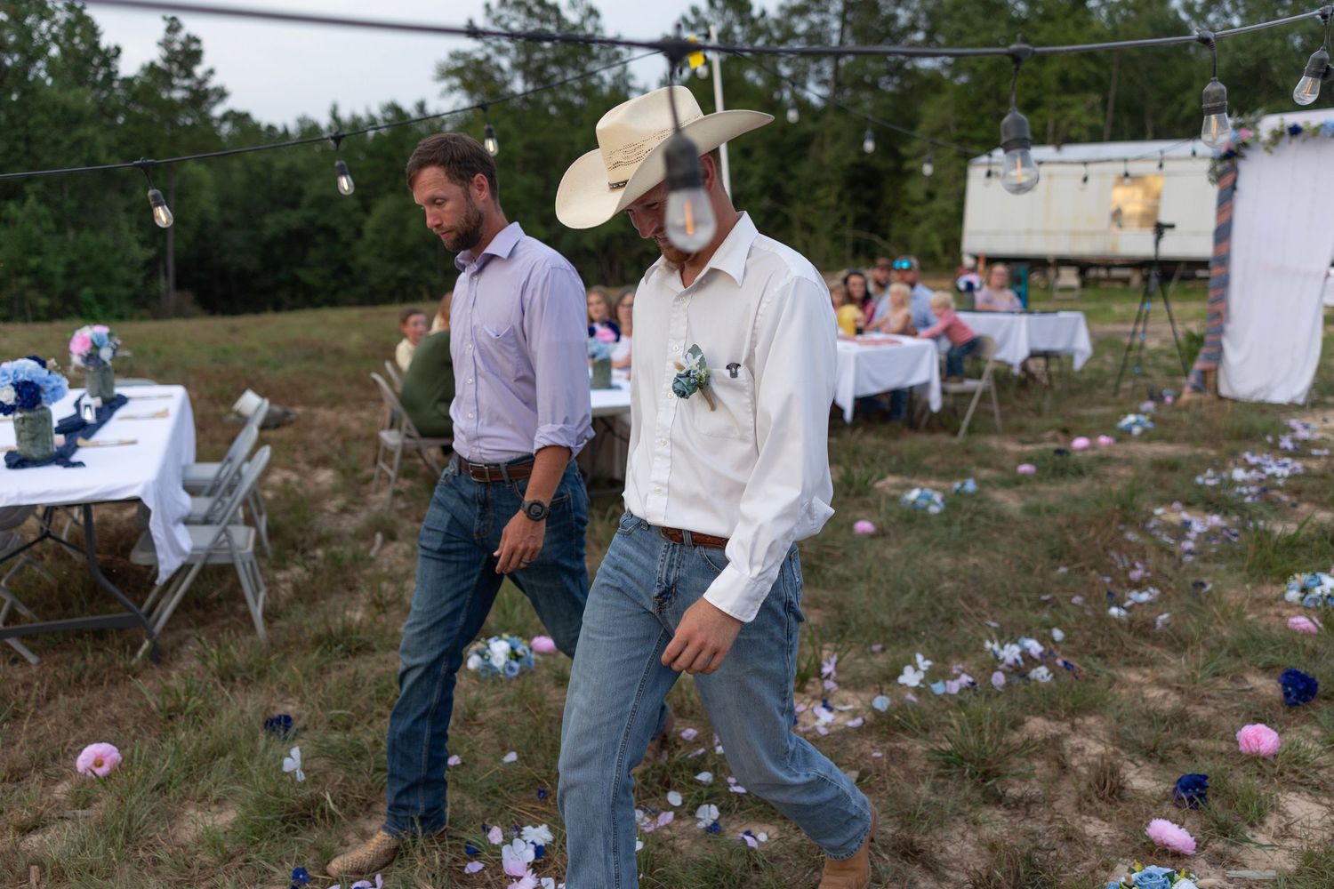 Two men walking through a littered outdoor party venue with string lights, one wearing a white cowboy hat.