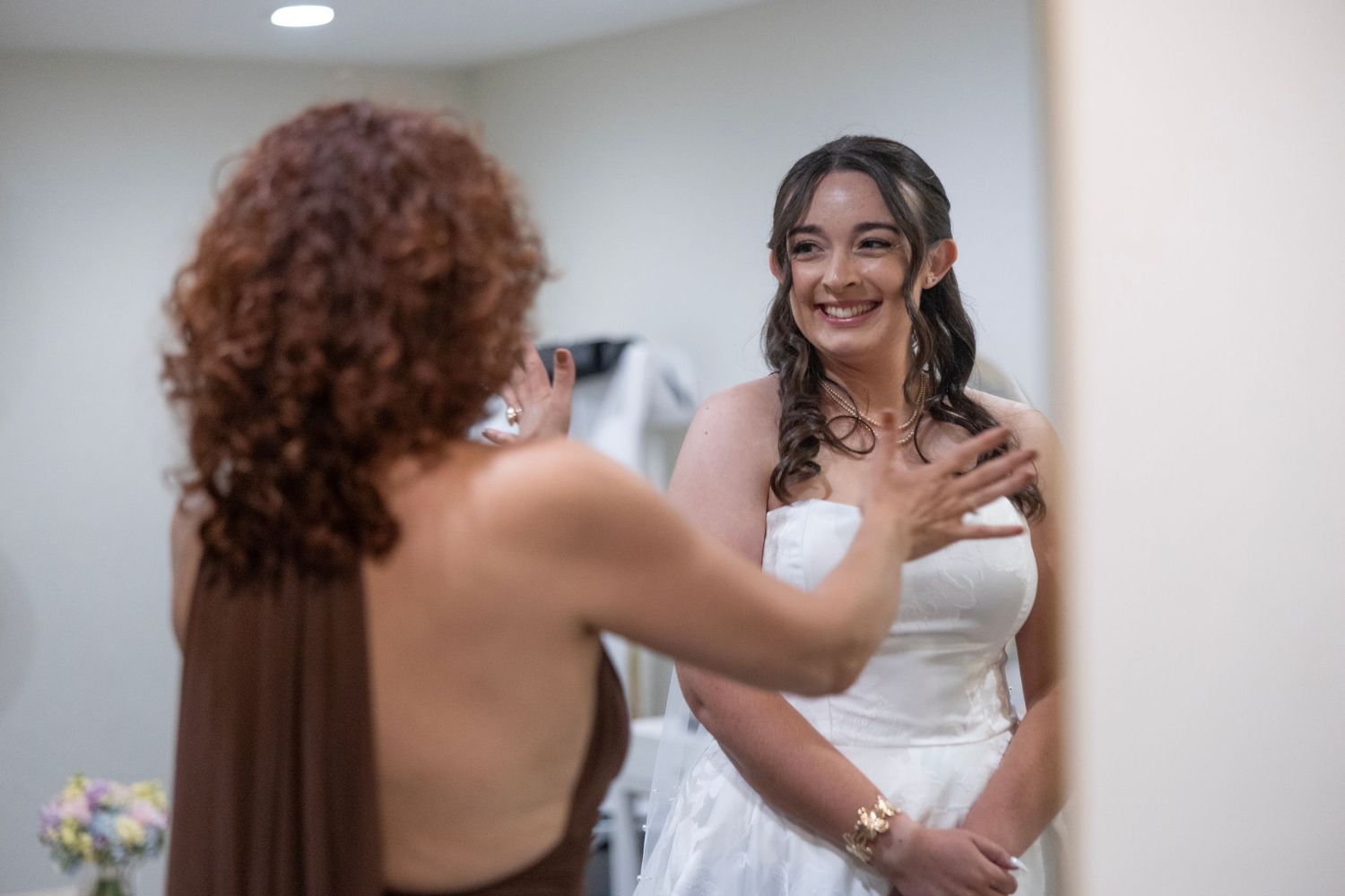 Bride in white wedding dress smiling at mirror while bridesmaid in brown dress helps her prepare for the ceremony.