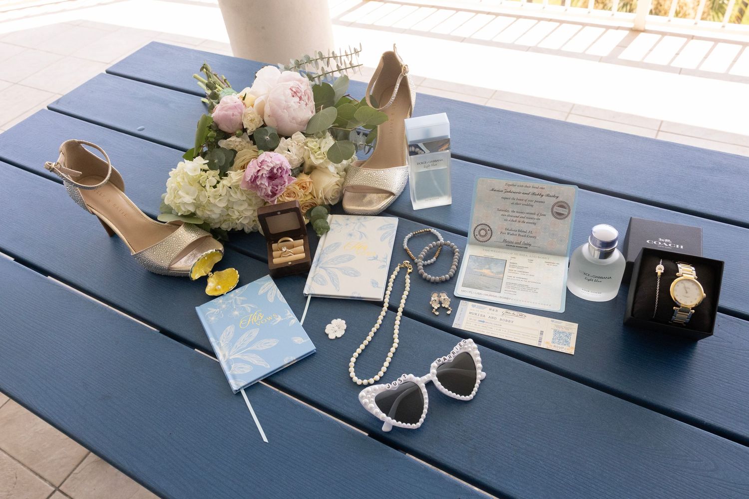 Wedding flat lay with bridal bouquet, heels, pearl necklace, sunglasses, invitations, watch, and accessories on blue table.