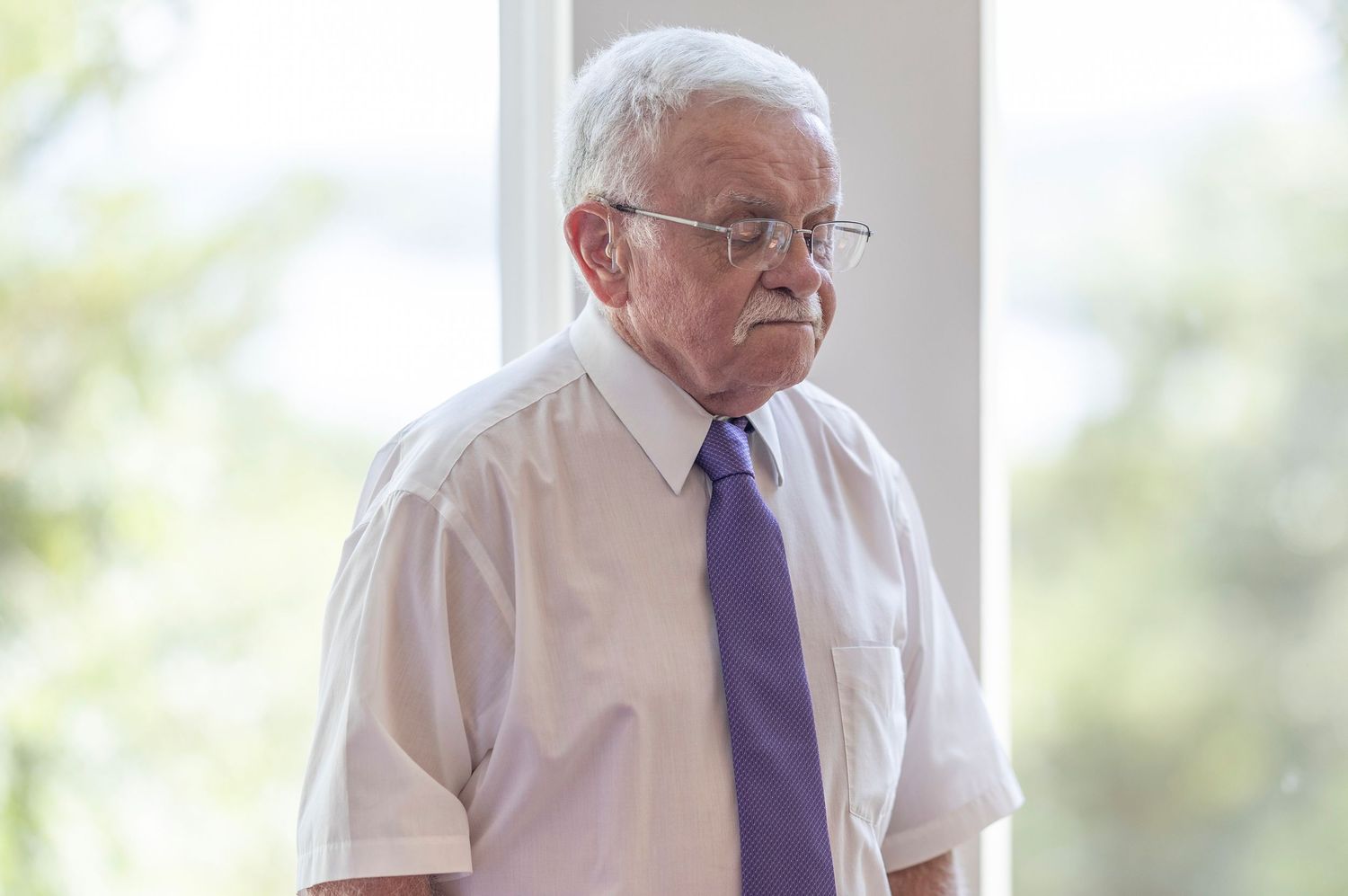 Elderly man with white hair wearing a white shirt and purple tie, standing indoors with a thoughtful expression.