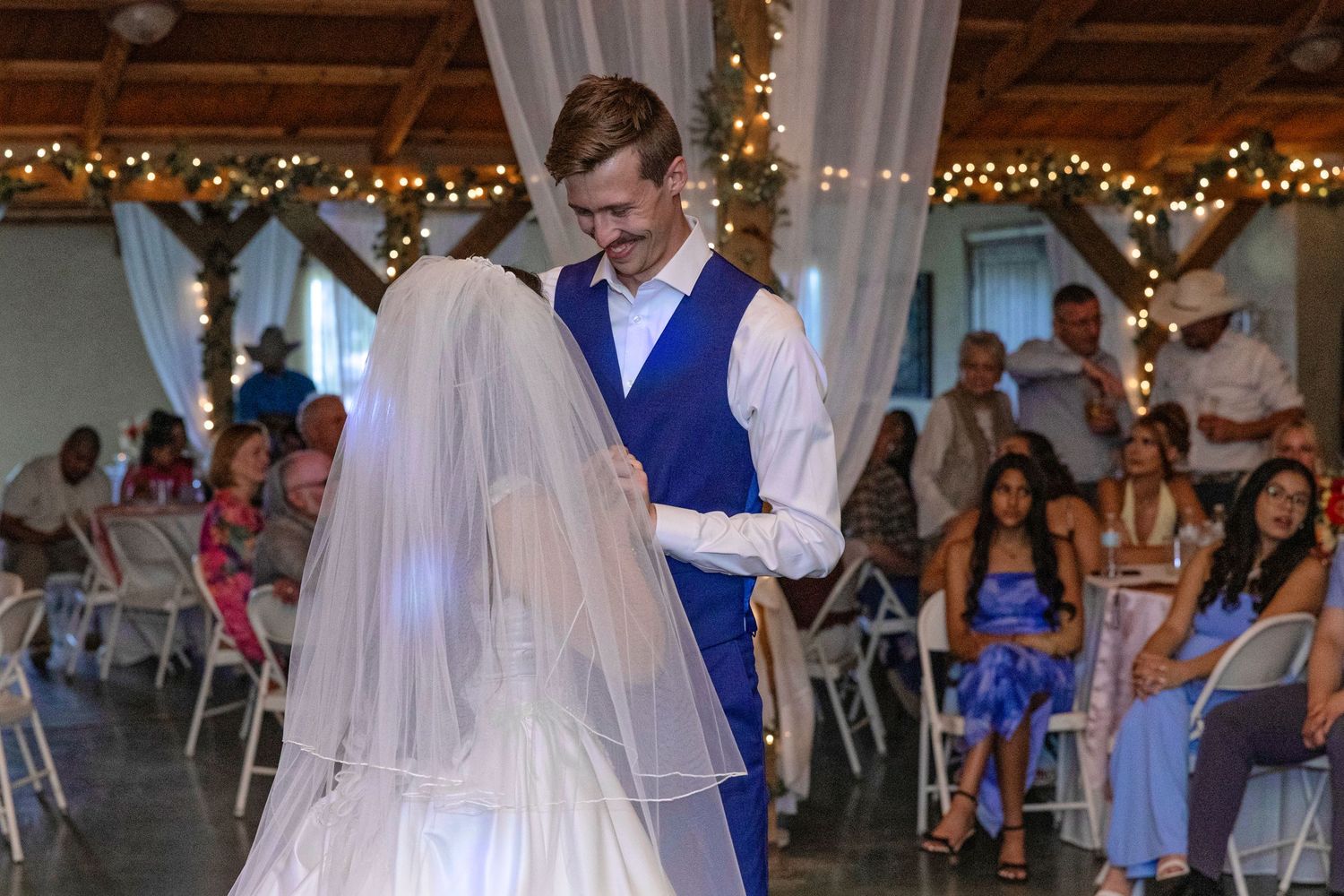 Bride and groom share first dance at elegant wedding reception with string lights and seated guests.
