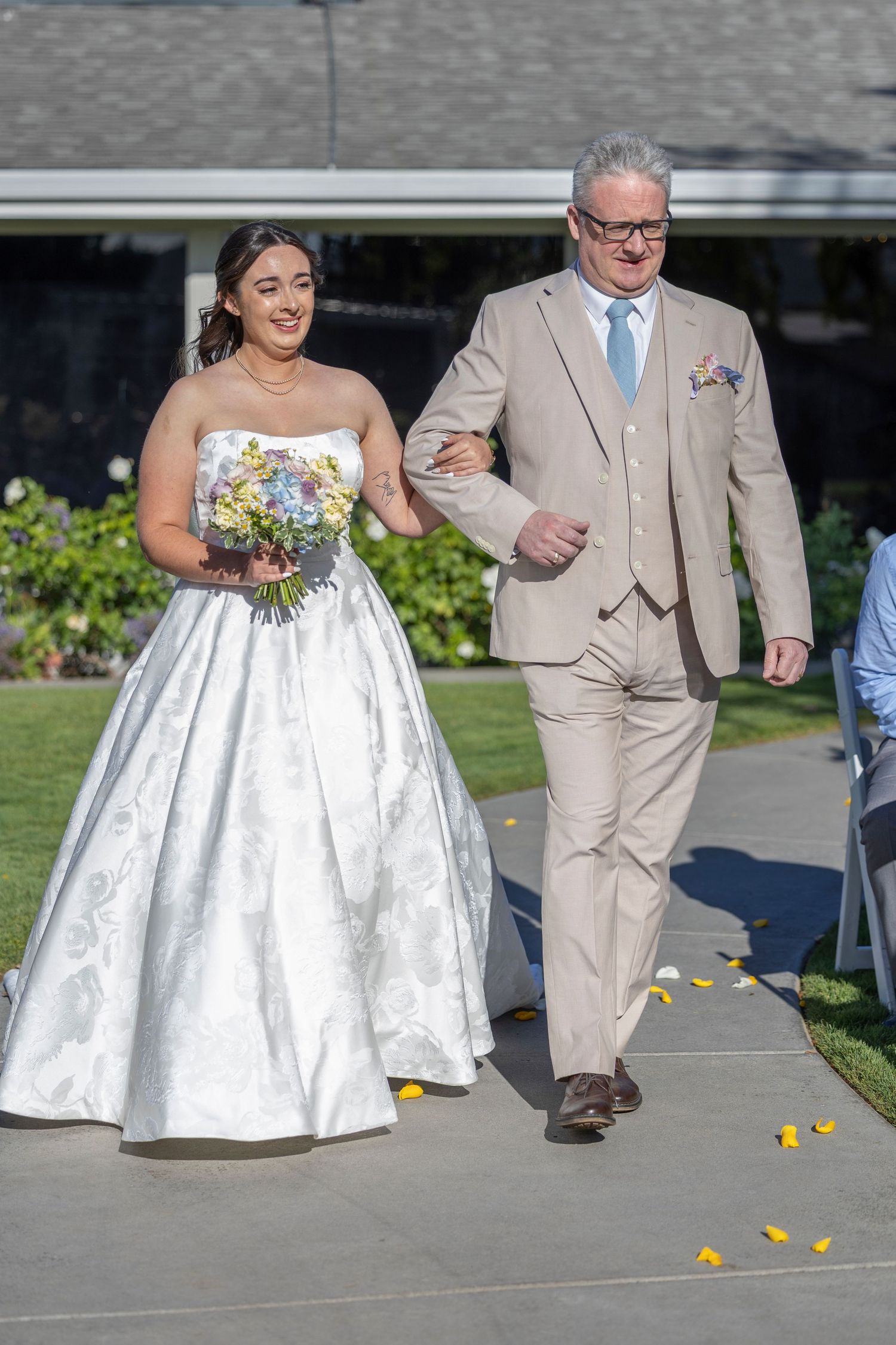 Bride in white strapless gown holding floral bouquet walks down aisle with man in tan suit at outdoor wedding ceremony.