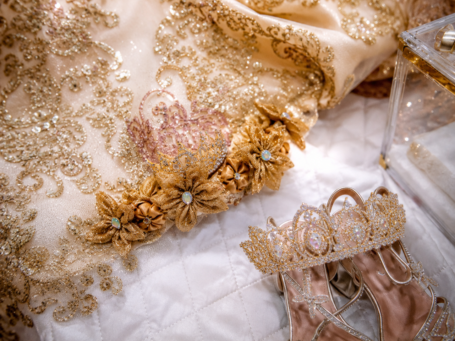 Gold embroidered bridal gown with floral sash, crystal crown, and elegant heeled sandals on white quilted surface.