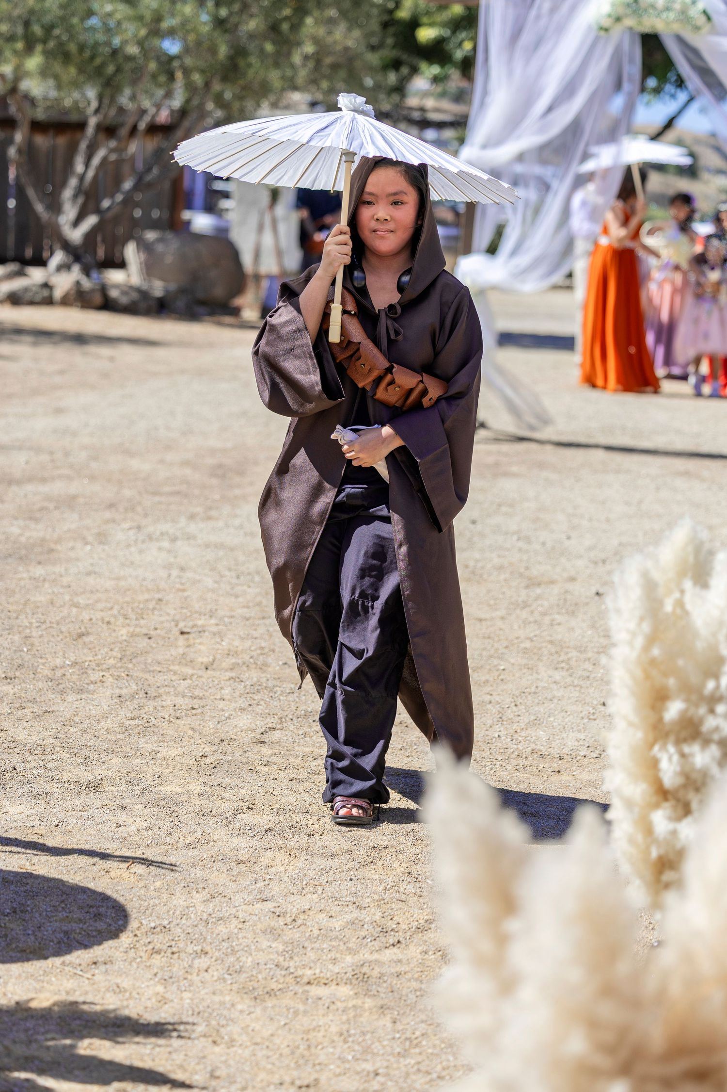 Young girl in dark hooded robe holding white parasol umbrella walks along sandy path at outdoor event.