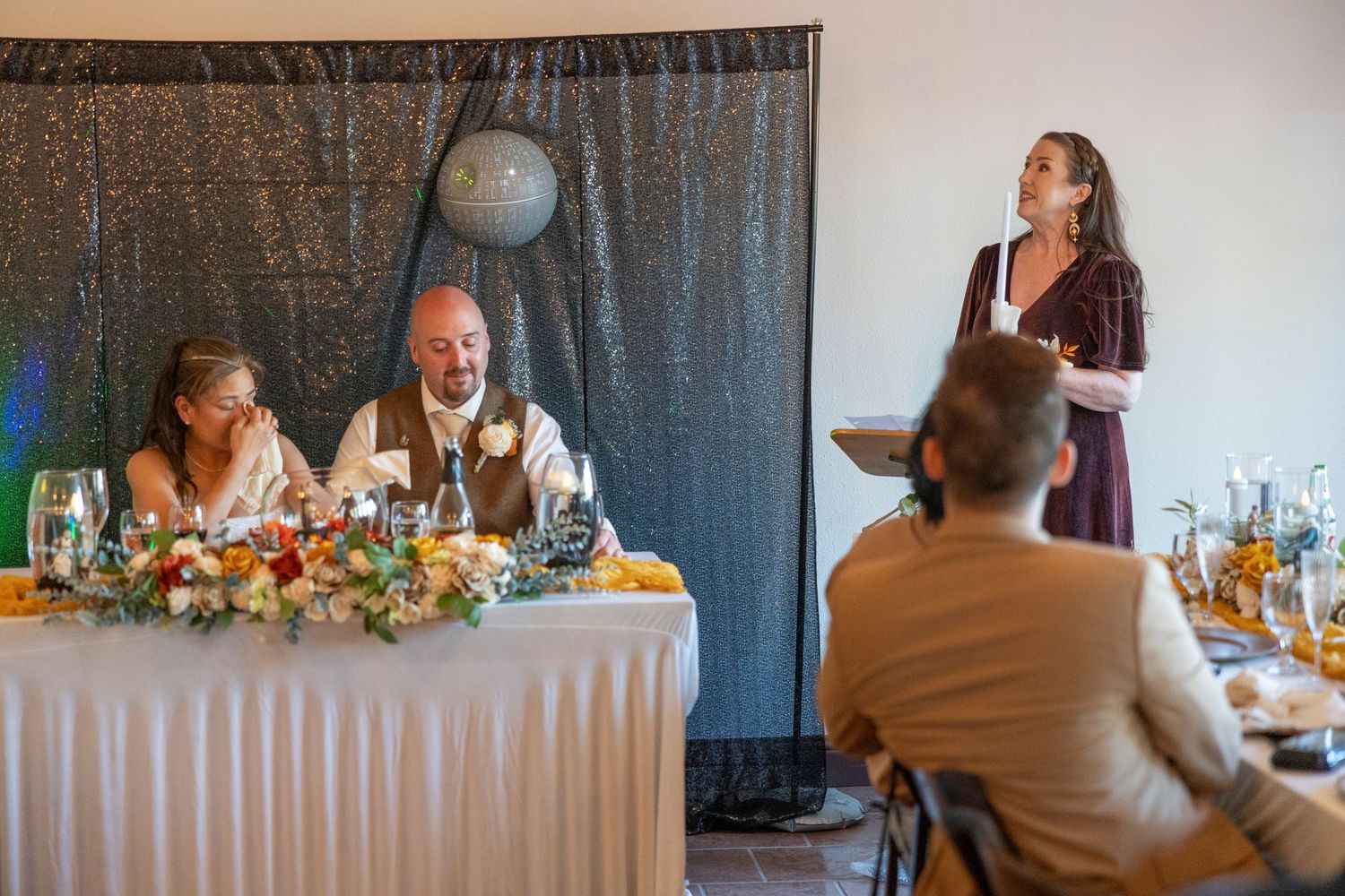 Woman in burgundy dress gives a toast at a wedding reception while the bride and groom smile at the head table.