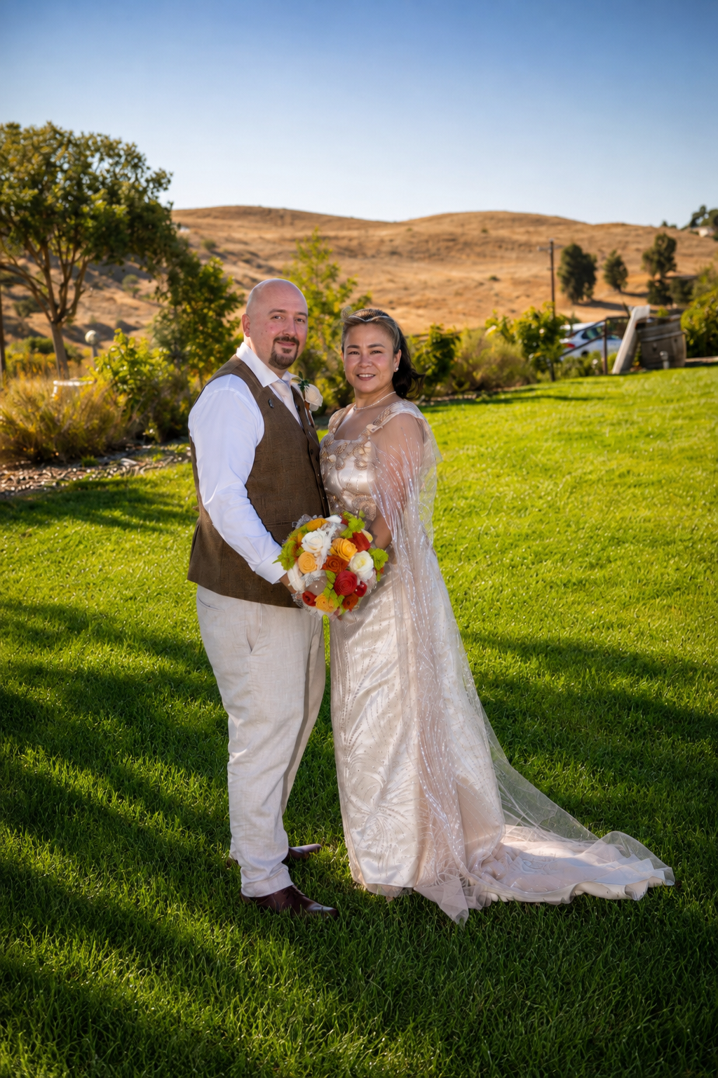 A smiling bride and groom pose on green grass with golden hills behind them on their wedding day.