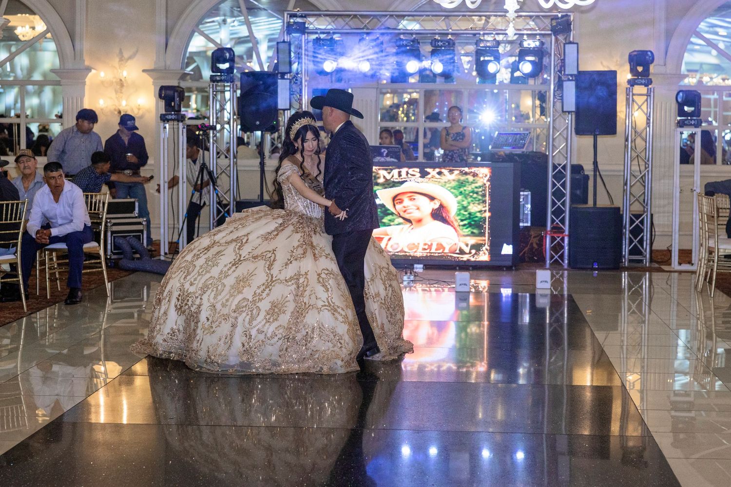 A young woman in a gold quinceañera gown dances with a man in a cowboy hat on a shiny dance floor at a festive celebration.