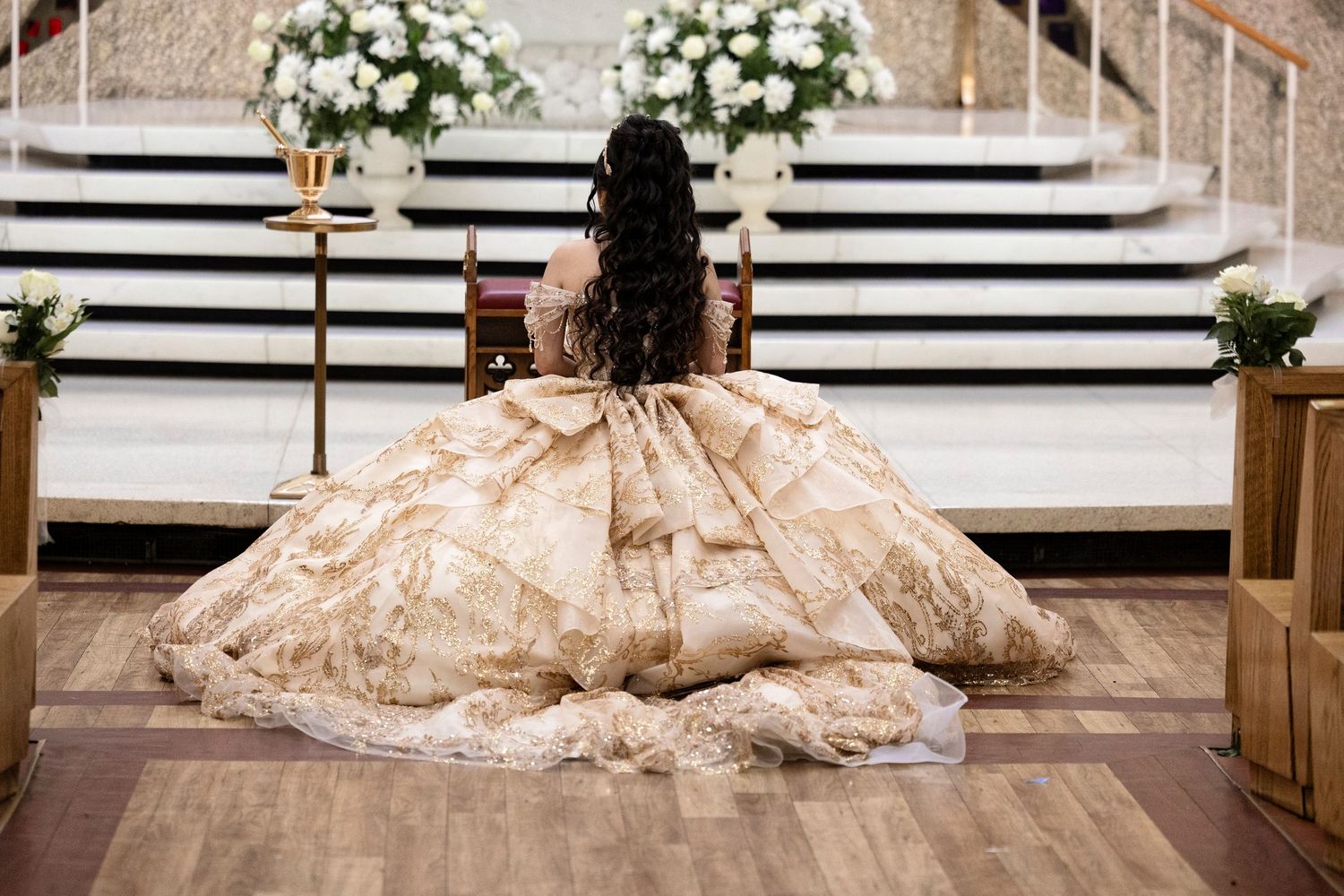 Young woman in ornate gold ball gown kneels at church altar during quinceañera ceremony.