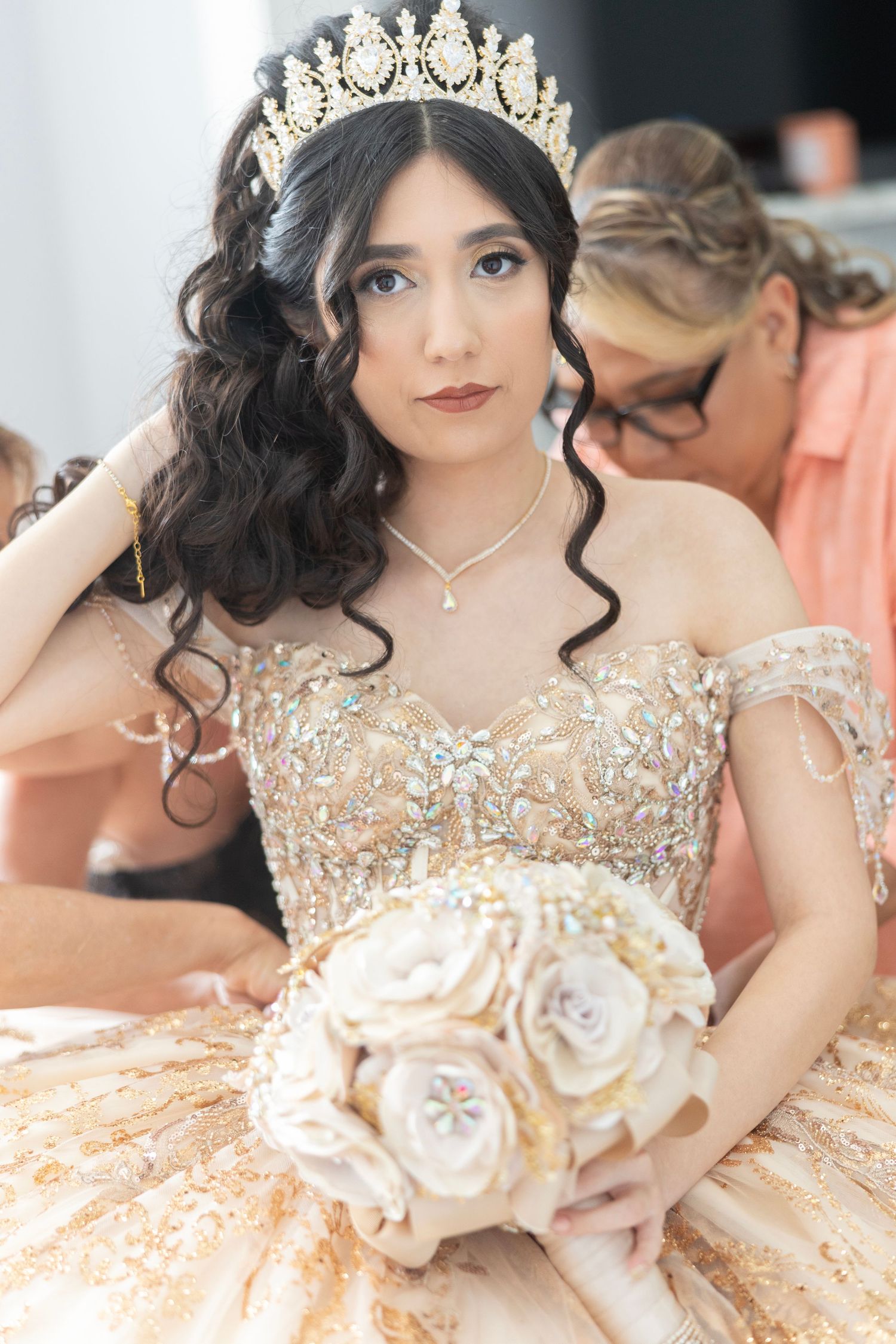 Young woman in a gold quinceañera gown with a jeweled crown holds a white floral bouquet, posing elegantly.