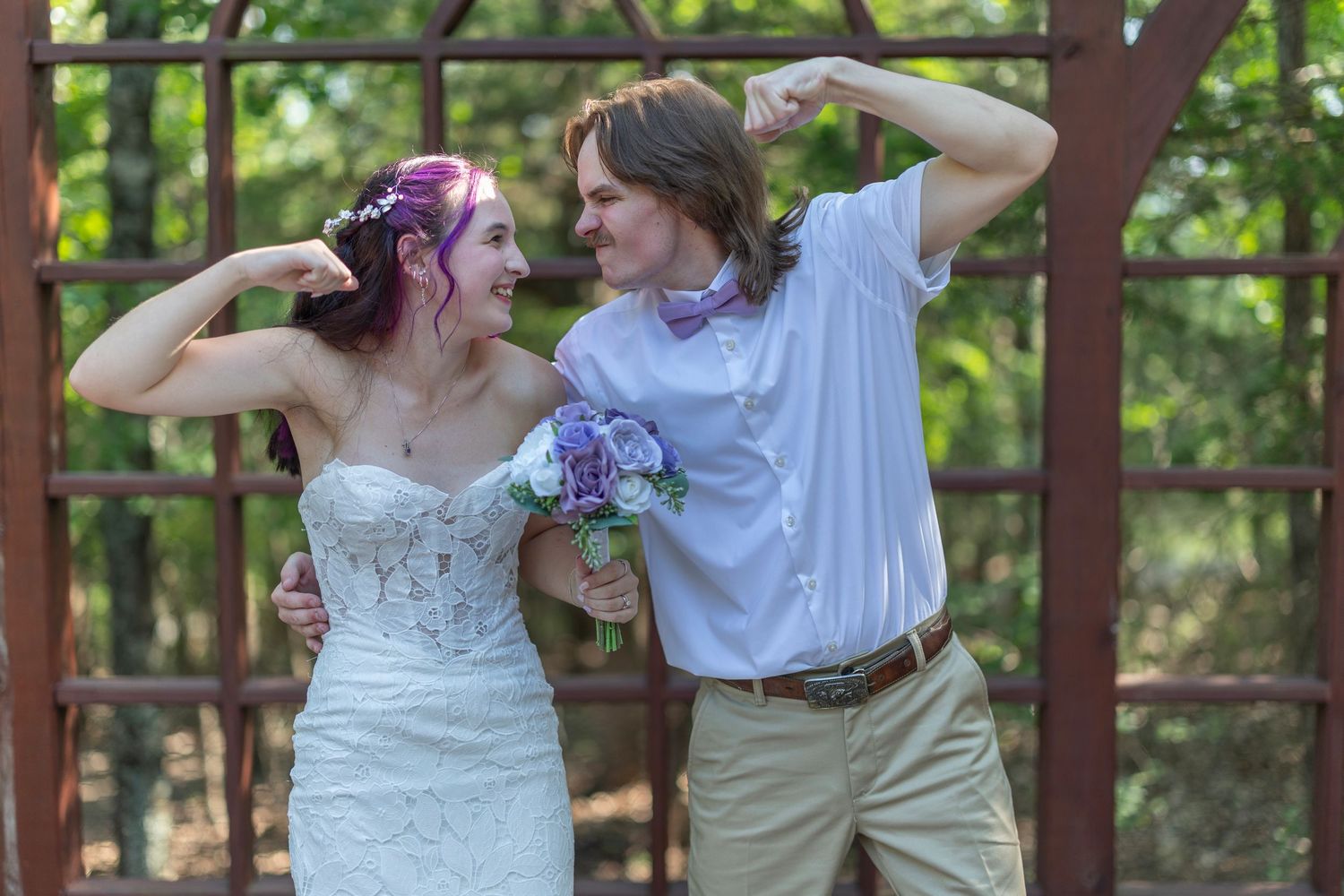 Bride and groom flexing muscles and laughing together outdoors, bride holding purple bouquet in white lace wedding dress.