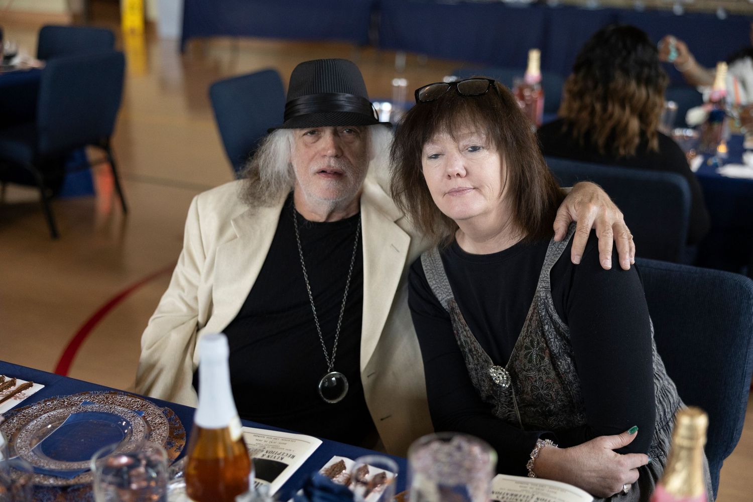 Two adults pose together at a banquet table, man in white blazer with arm around woman in black outfit.