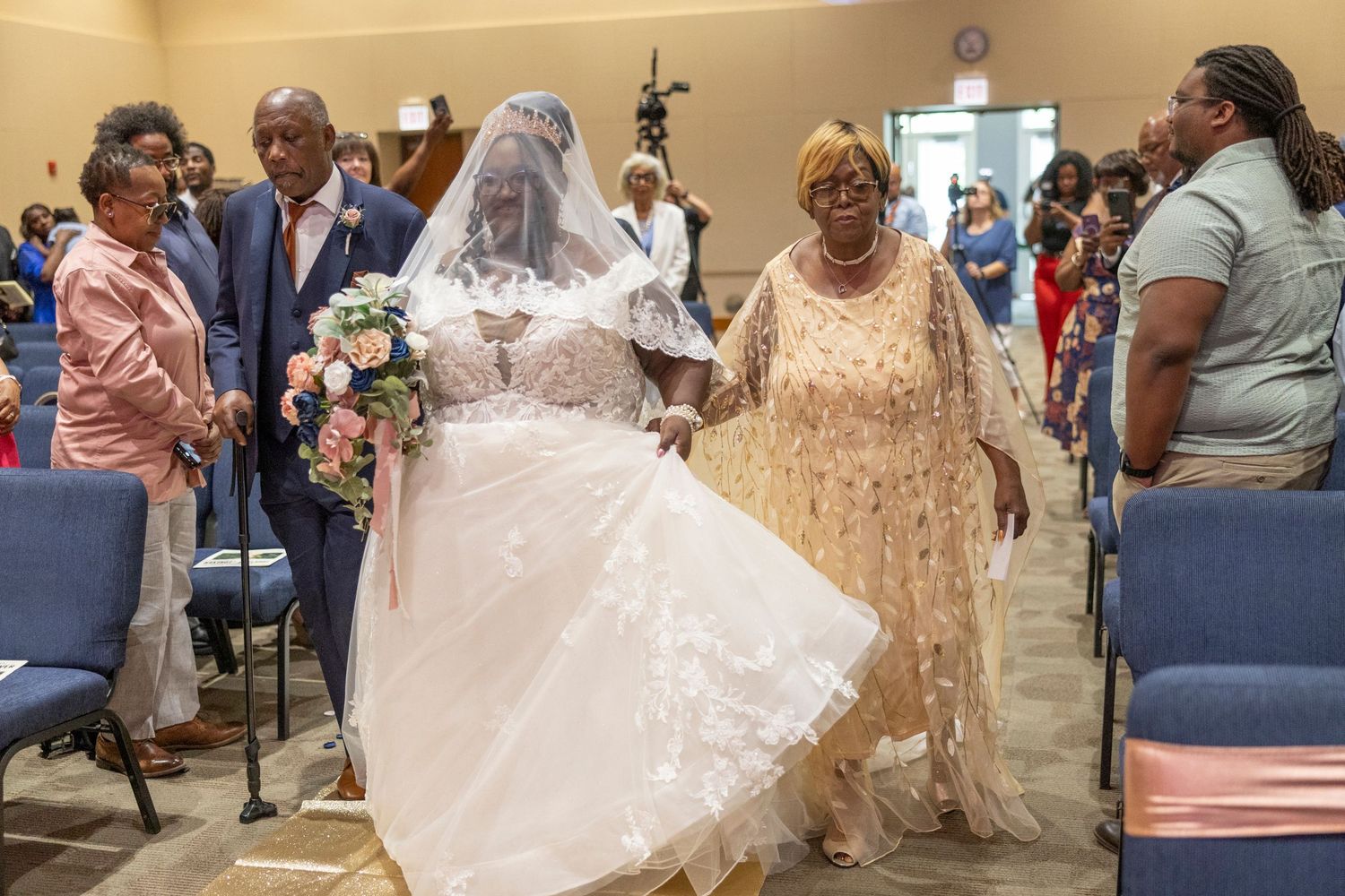Bride in white gown and veil walks down the aisle with family at an indoor wedding ceremony.