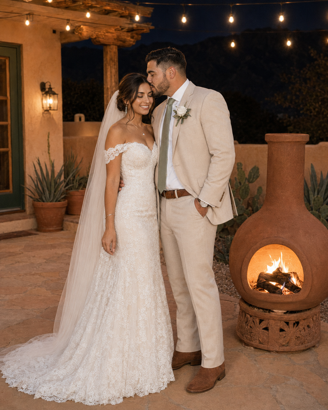 Couple in wedding attire poses by a glowing chiminea outdoors at night with string lights.