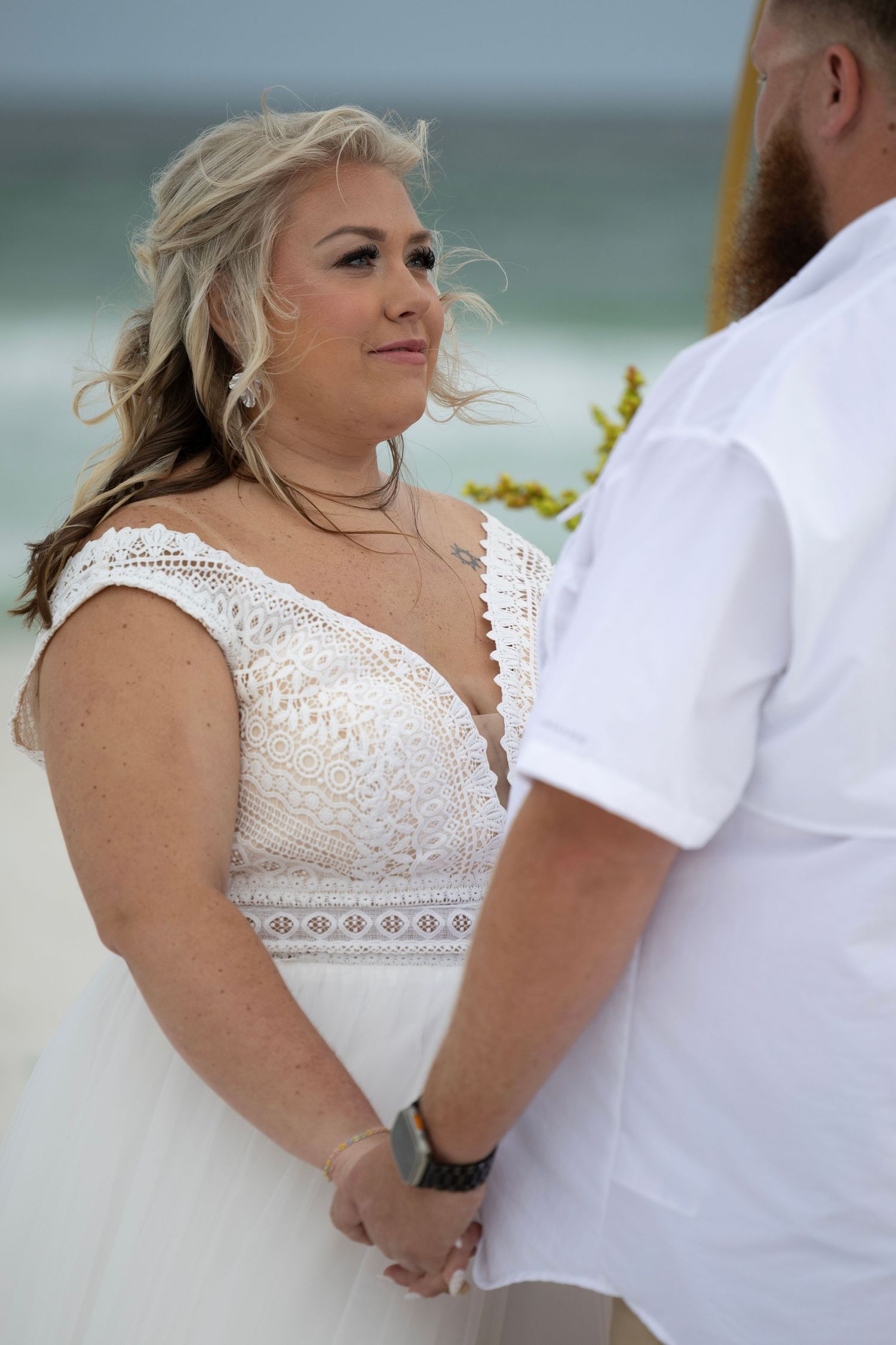 Bride in white lace dress holds hands with groom during romantic beach wedding ceremony.