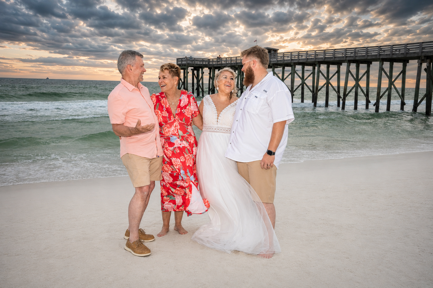 Couple in wedding attire poses with family on sandy beach near pier at sunset.