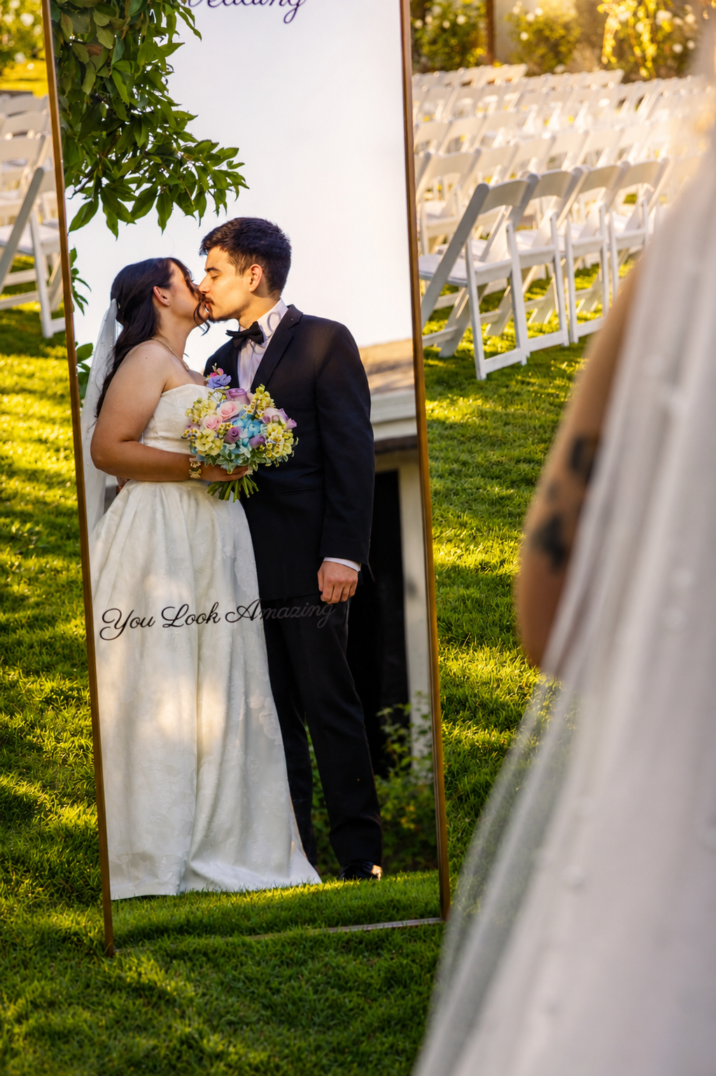 Bride and groom sharing a kiss reflected in a mirror at an outdoor wedding ceremony with white chairs on green lawn.