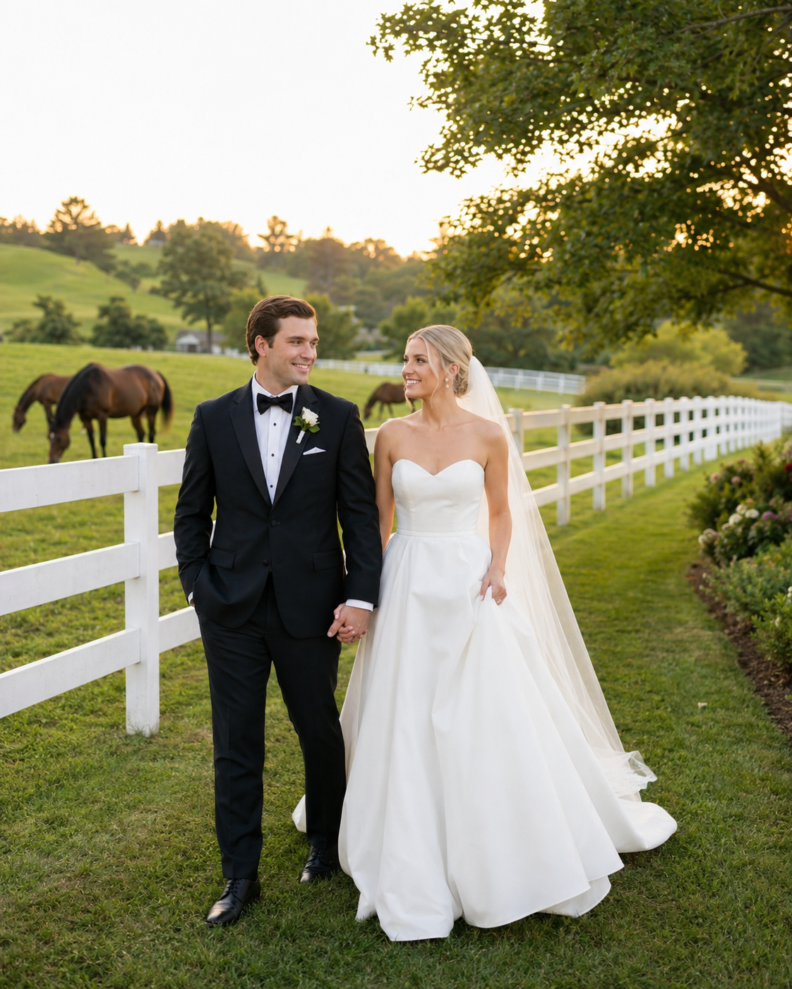 Bride and groom holding hands at sunset on a lush green farm with white fence and horses in background.