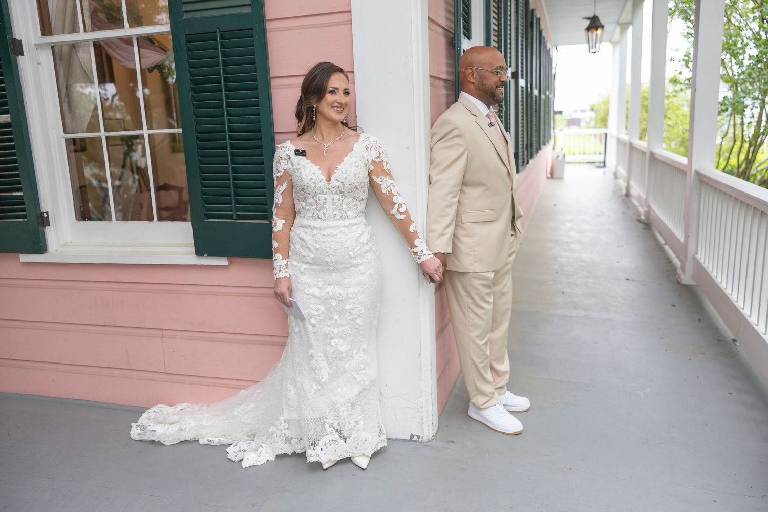 A couple shares a sweet first look moment on a covered porch of a pink historic building with green shutters.