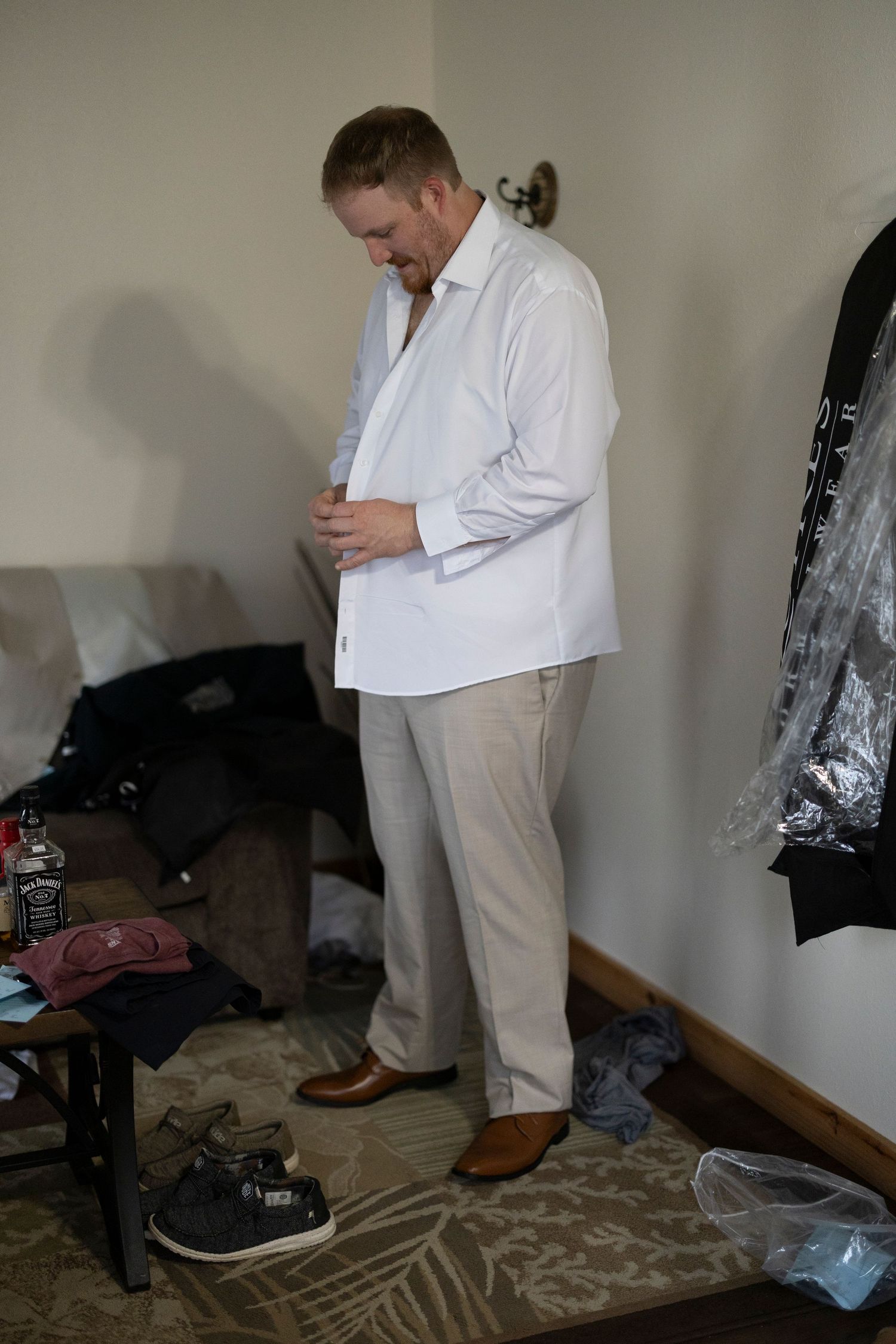 Someone in formal attire smiles while getting dressed in a room with scattered clothes and shoes on the floor.