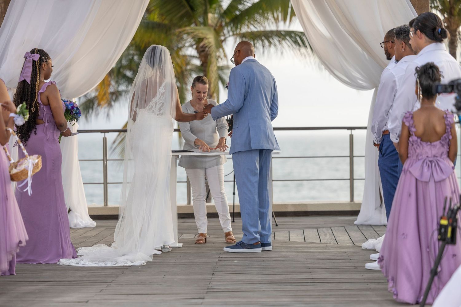 A wedding party in lavender dresses gathers on a covered outdoor deck during a celebration.