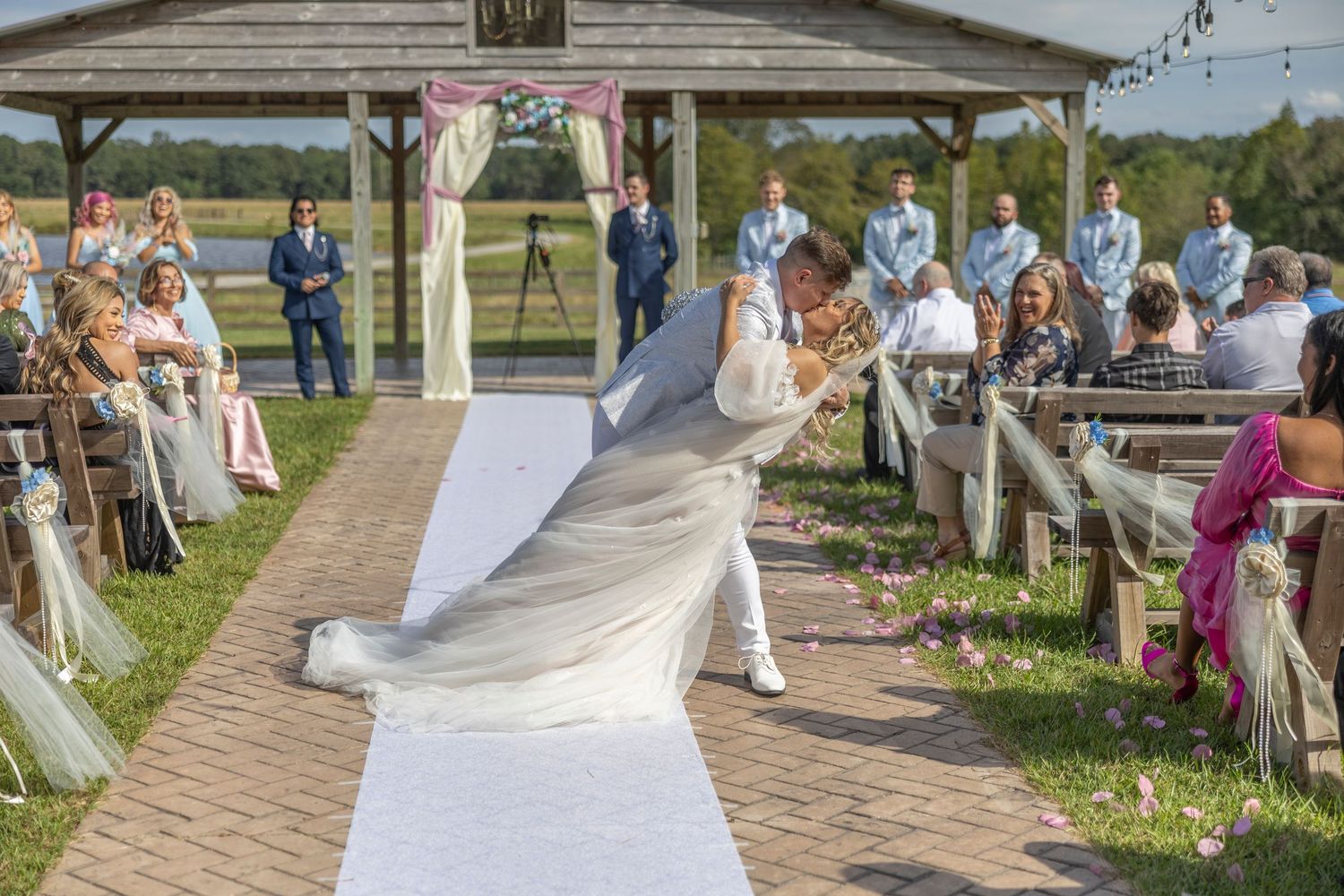 Wedding guests gather outdoors on a brick pathway during a sunny celebration with a gazebo in the background.