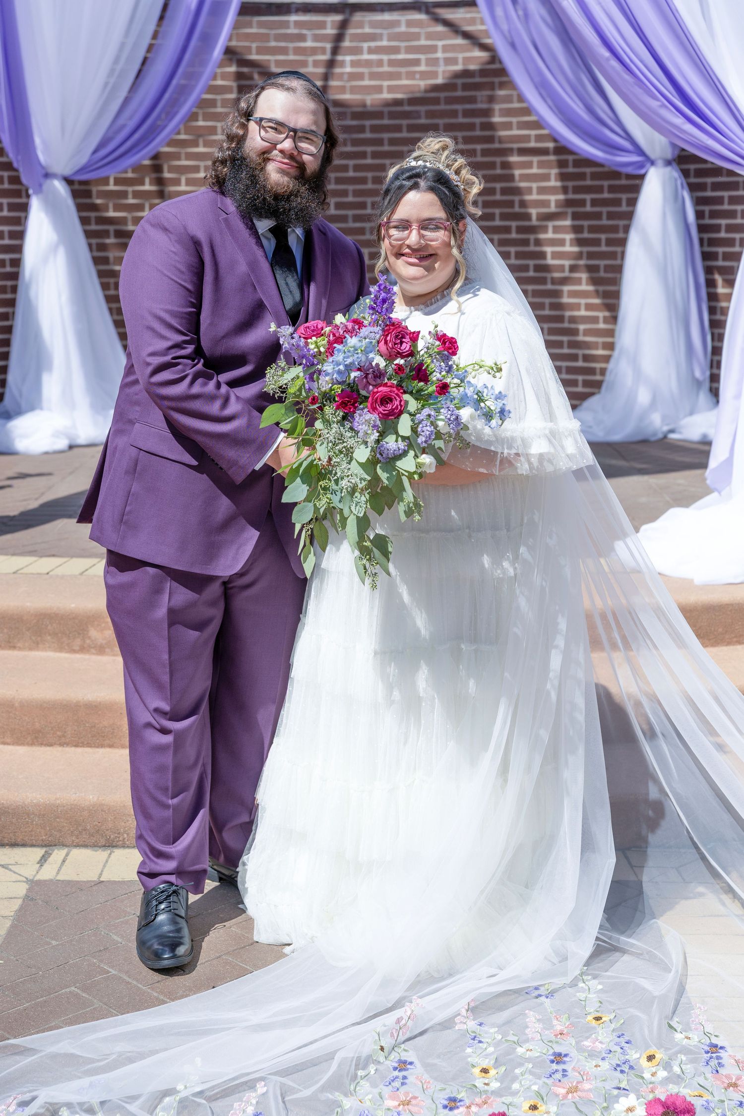 A couple in purple and white wedding attire share a joyful moment during their ceremony with floral decorations.