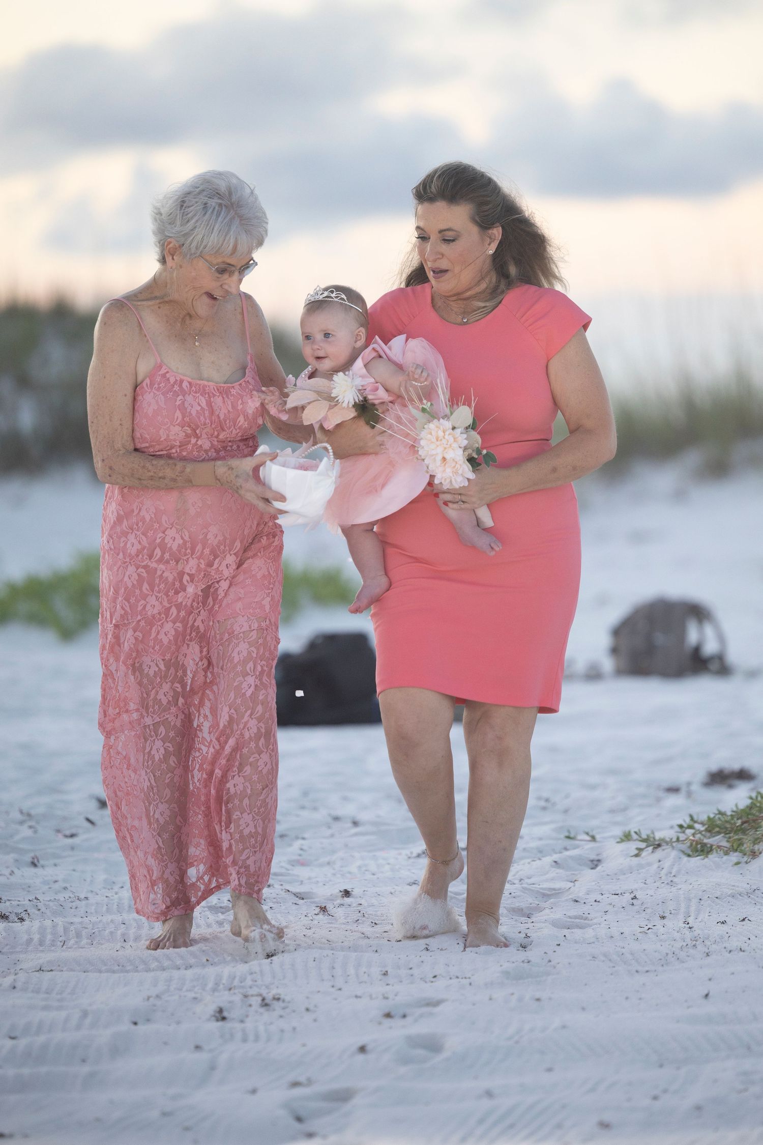 Three people in coral and pink outfits walk together on a sandy beach at sunset.