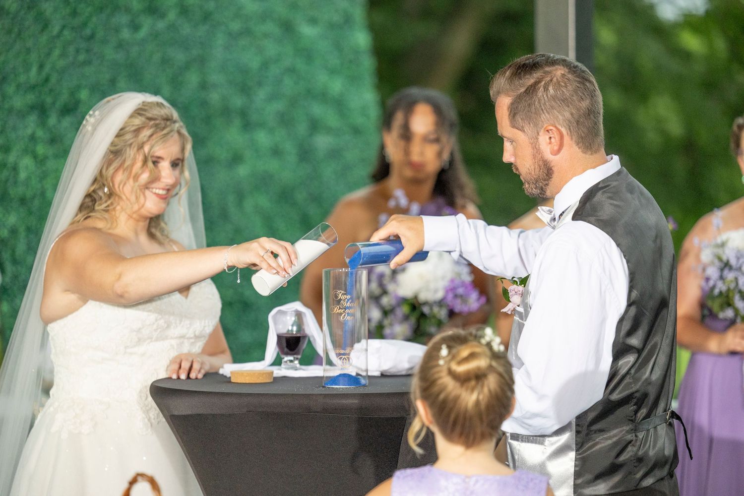 A wedding couple and guests enjoy a fun outdoor celebration with purple decor and natural greenery in the background.