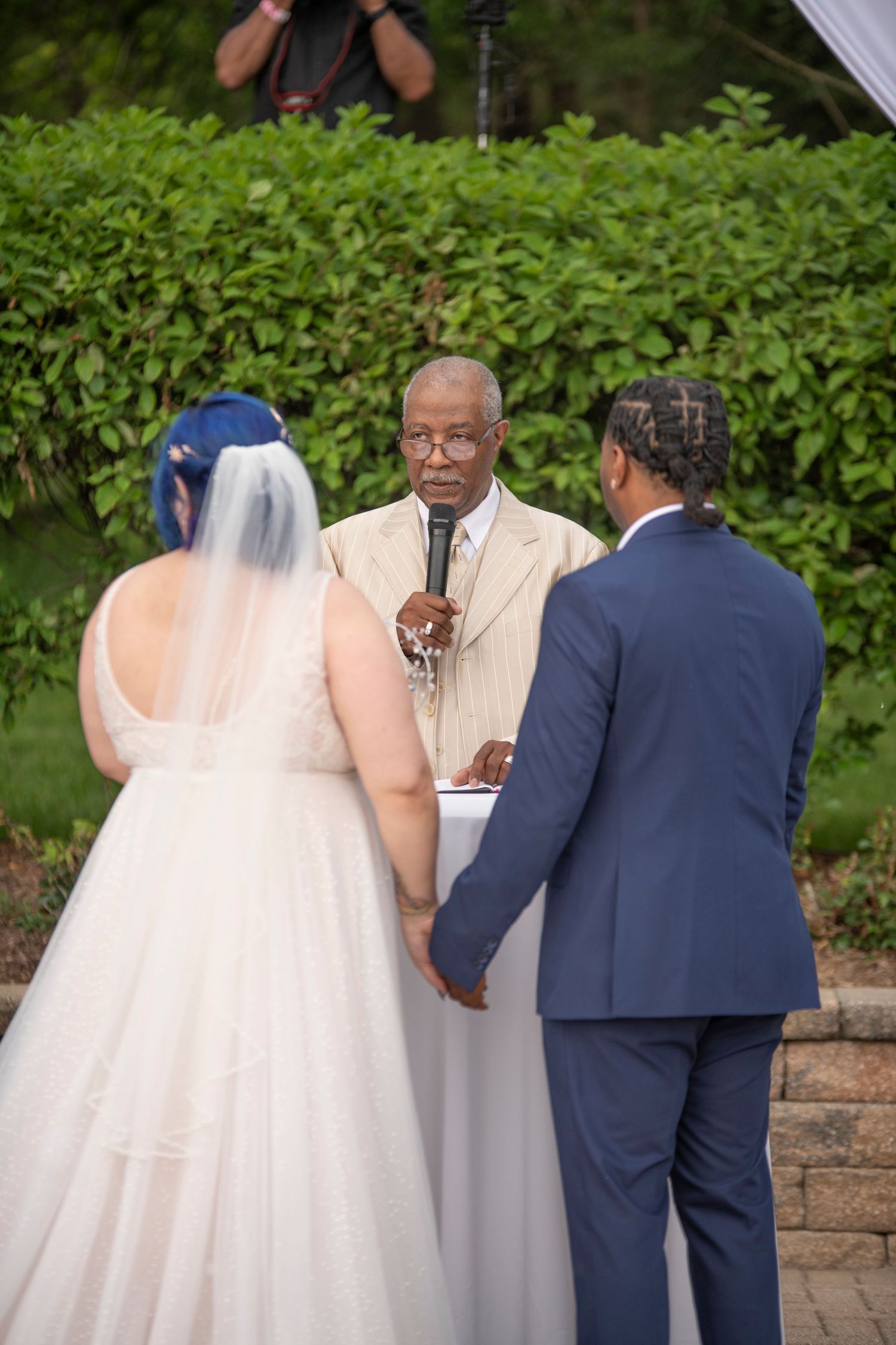 Wedding ceremony scene outdoors with couple and officiant standing together in front of green hedge backdrop.