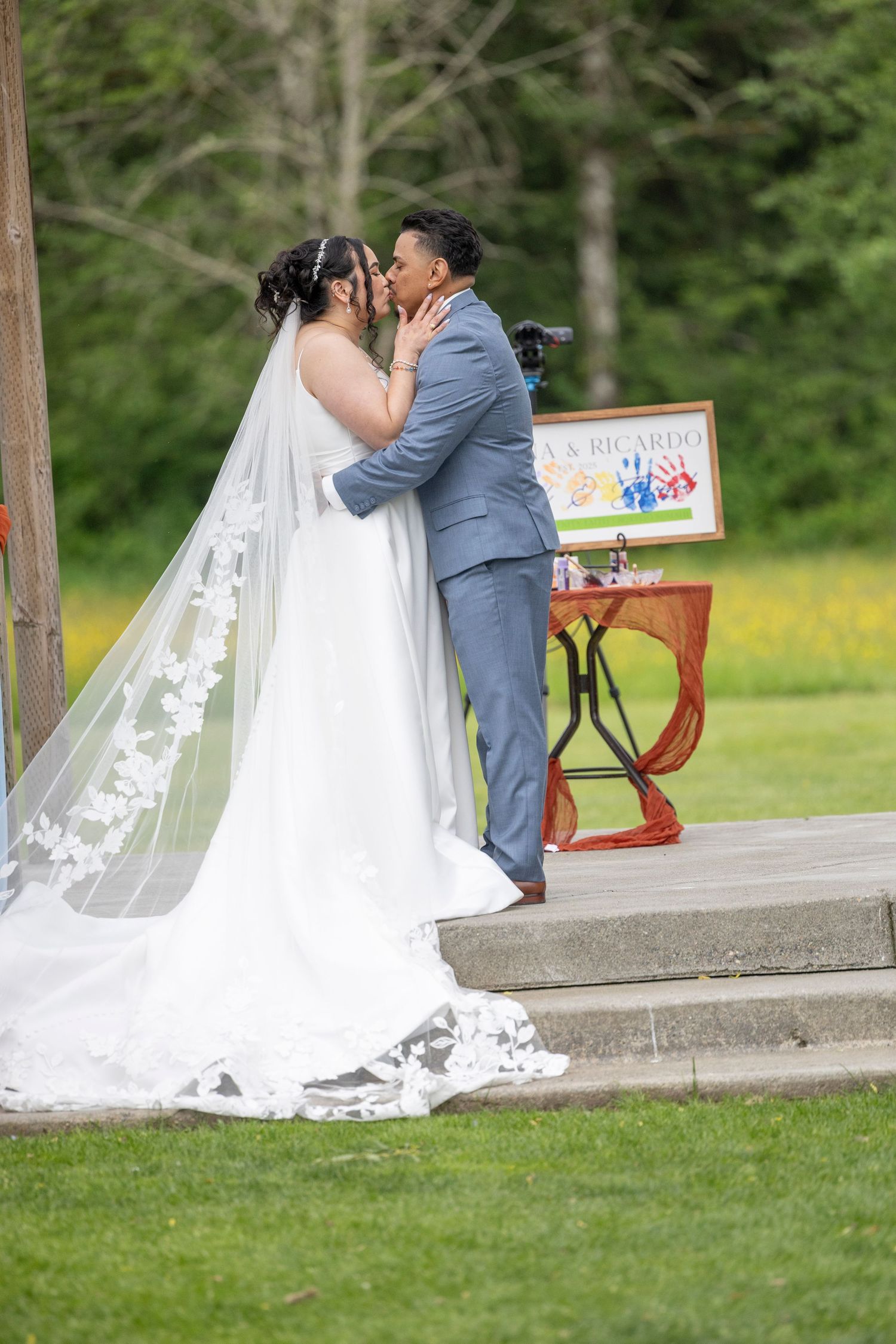 A romantic moment captured on a park bench as newlyweds share a tender kiss, with the bride's white lace gown flowing behind.
