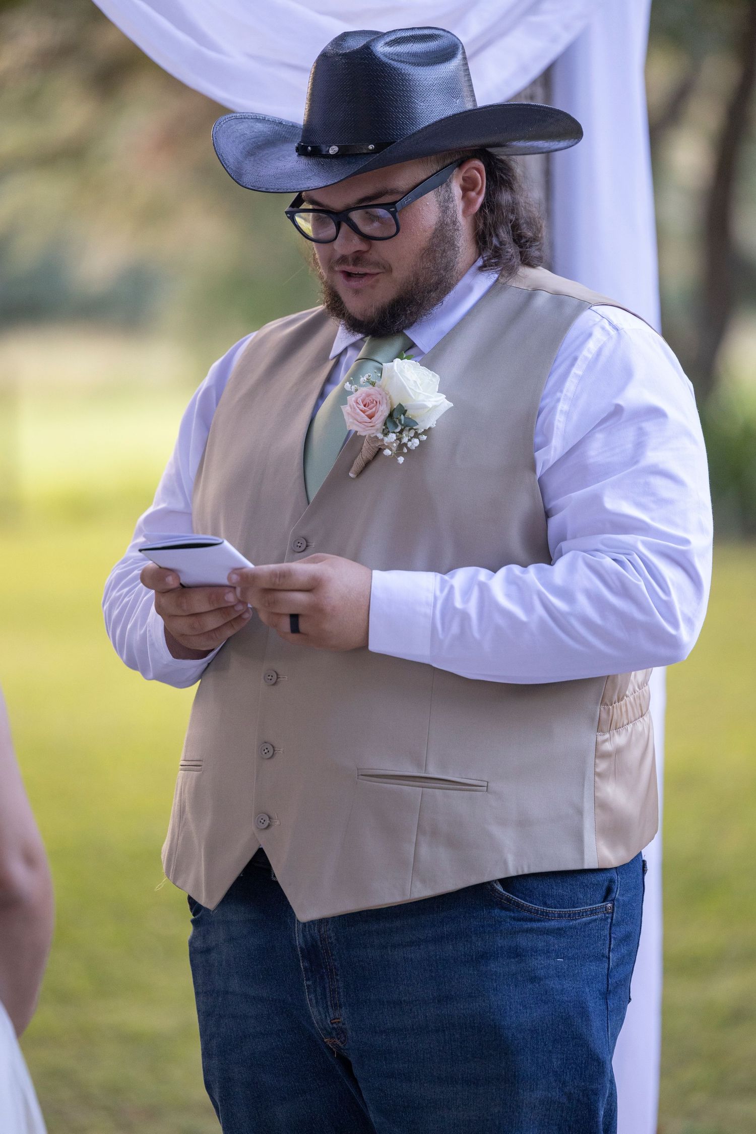 A groomsman in a cowboy hat, vest and white shirt reads from a phone during an outdoor wedding ceremony.