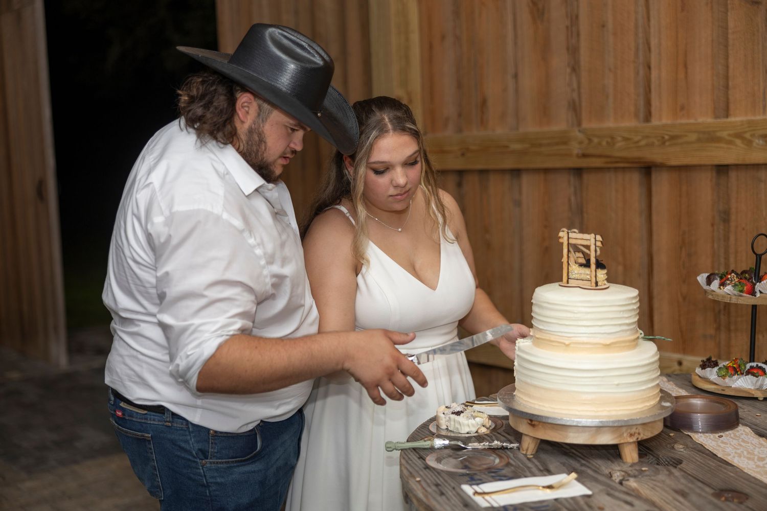 A couple in western attire cuts their white tiered wedding cake together at a rustic barn reception.