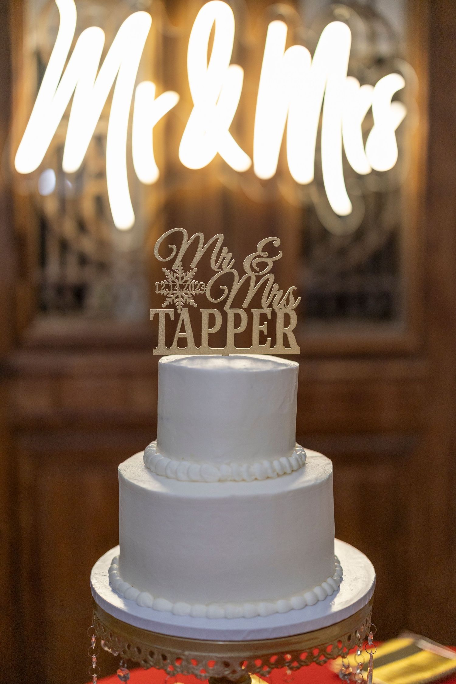 White wedding cake with elegant Mr & Mrs topper displayed on silver stand in warm lighting at reception venue.