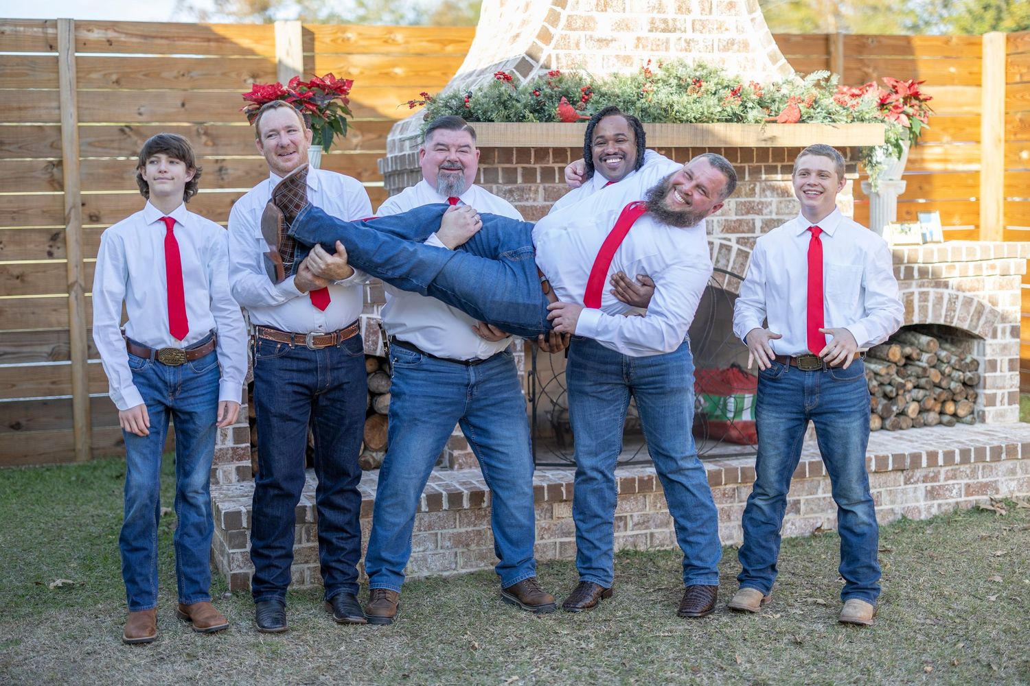 A playful group wearing matching red ties and jeans pose together outdoors in front of a wooden fence.