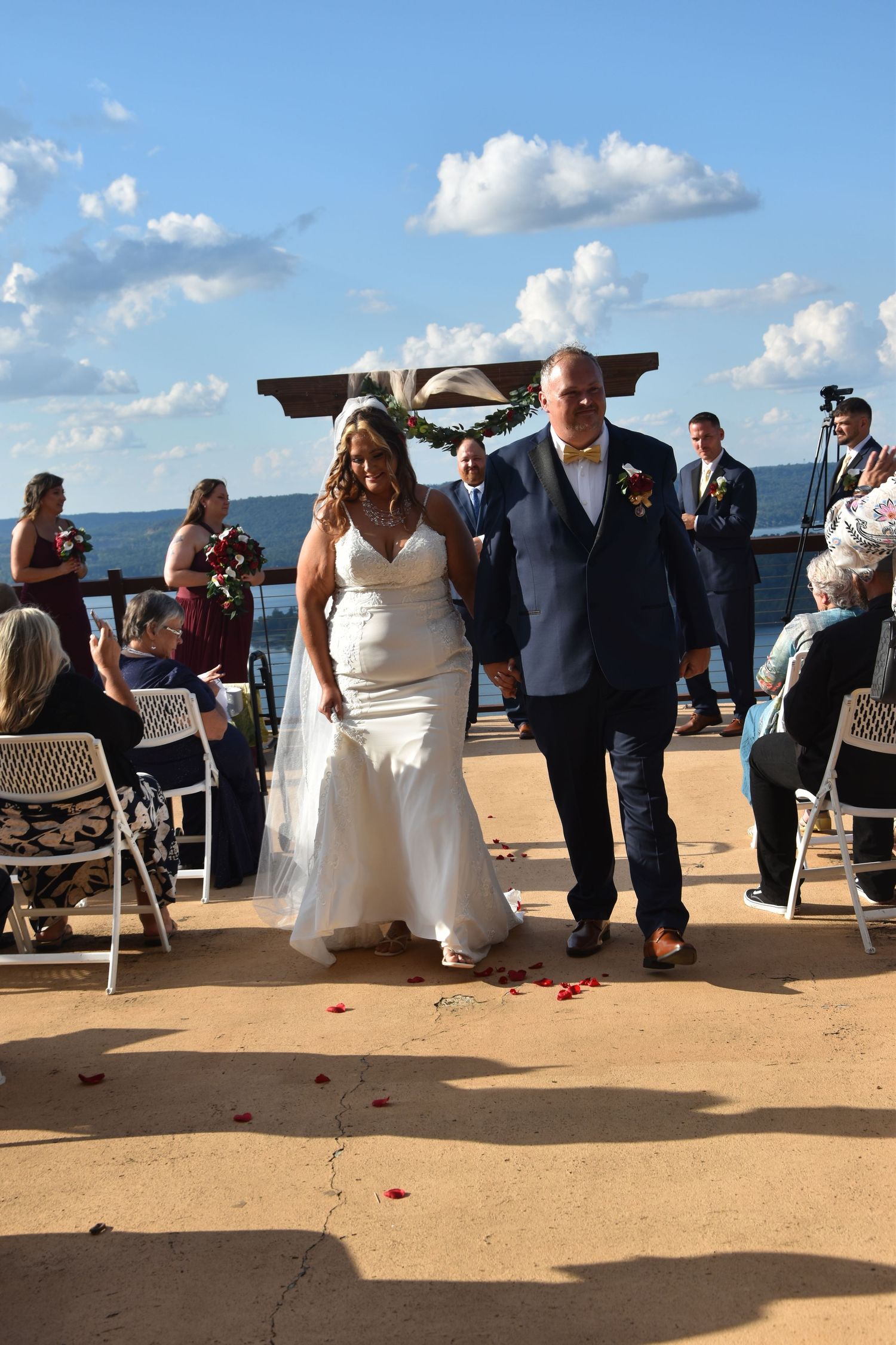 Wedding guests seated on white chairs at an outdoor beach ceremony with blue sky and clouds in the background.