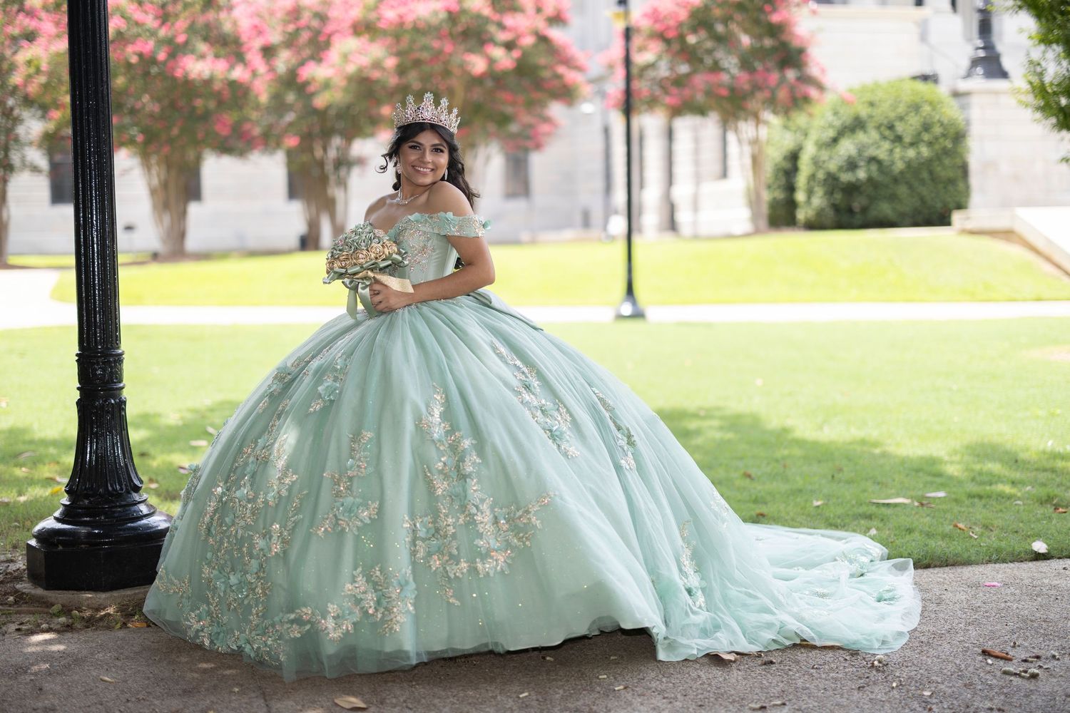 A person wearing a mint green ball gown with gold embellishments poses in a park setting during a photoshoot.