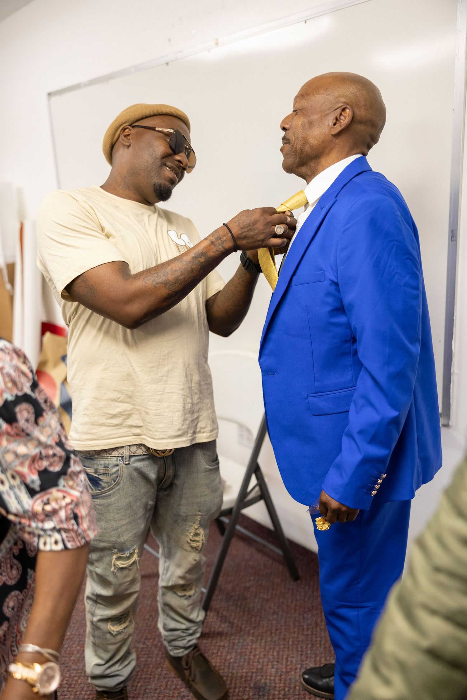 Man in beige hat adjusts yellow tie on man wearing vibrant blue suit indoors.