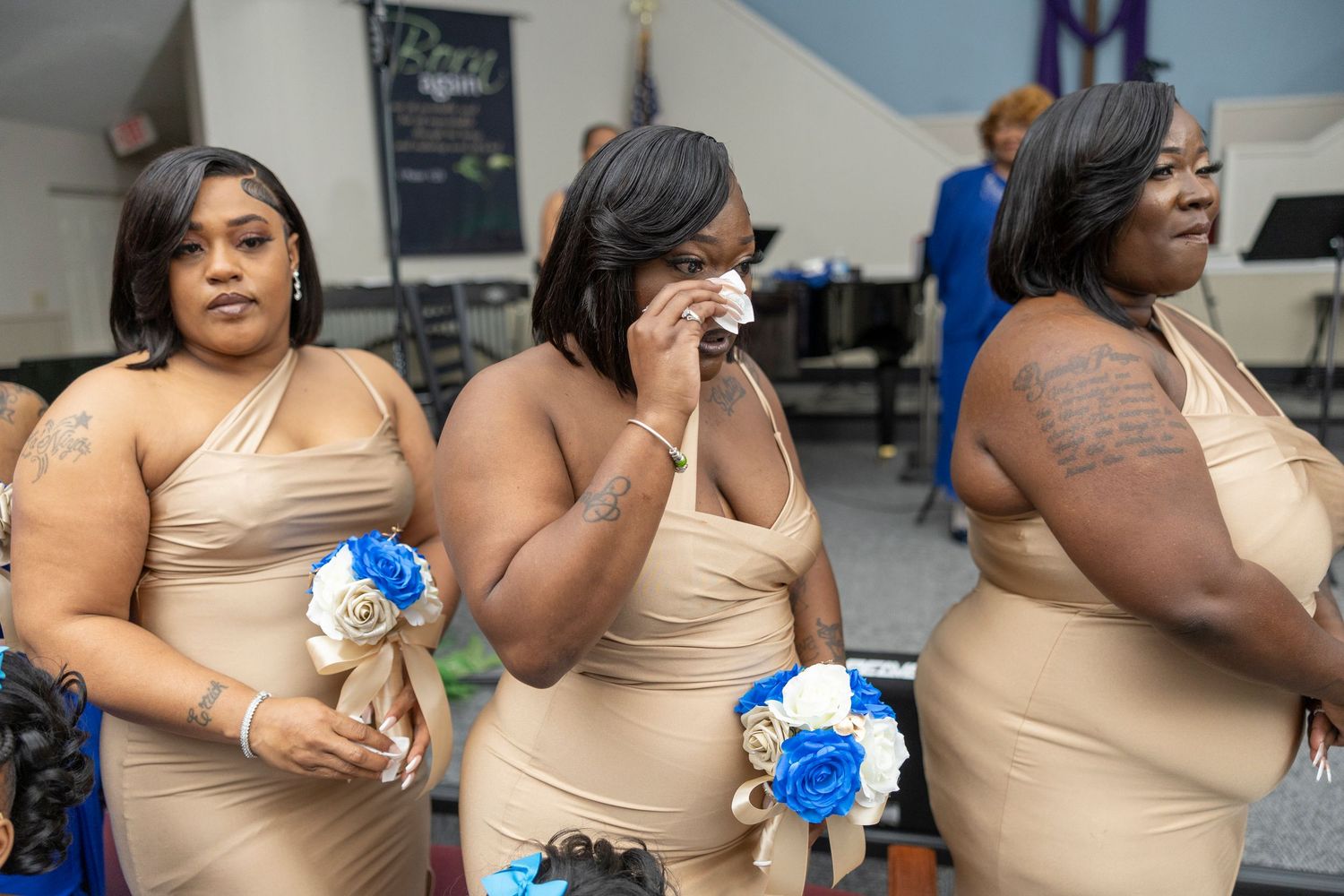 Three bridesmaids in tan dresses holding blue and white bouquets, one wiping tears at a wedding ceremony.
